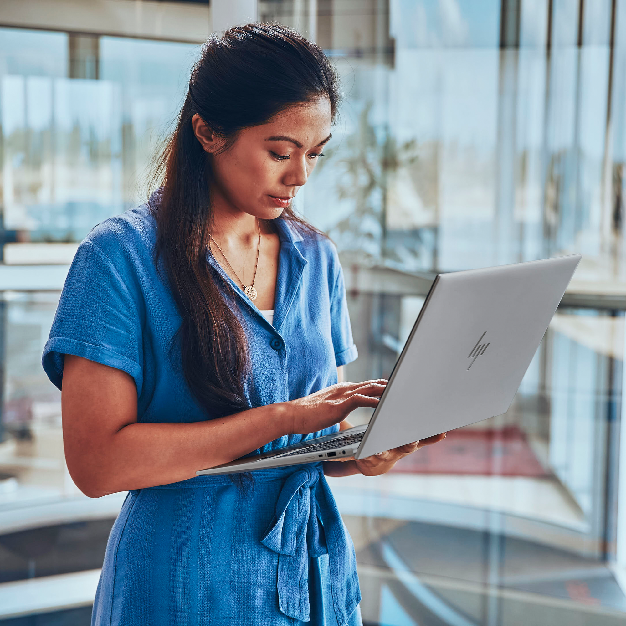 Frau in blauem Kleid, die einen silbernen Laptop benutzt. Sie schaut auf den Bildschirm. Hintergrund ist ein verschwommenes Büro.