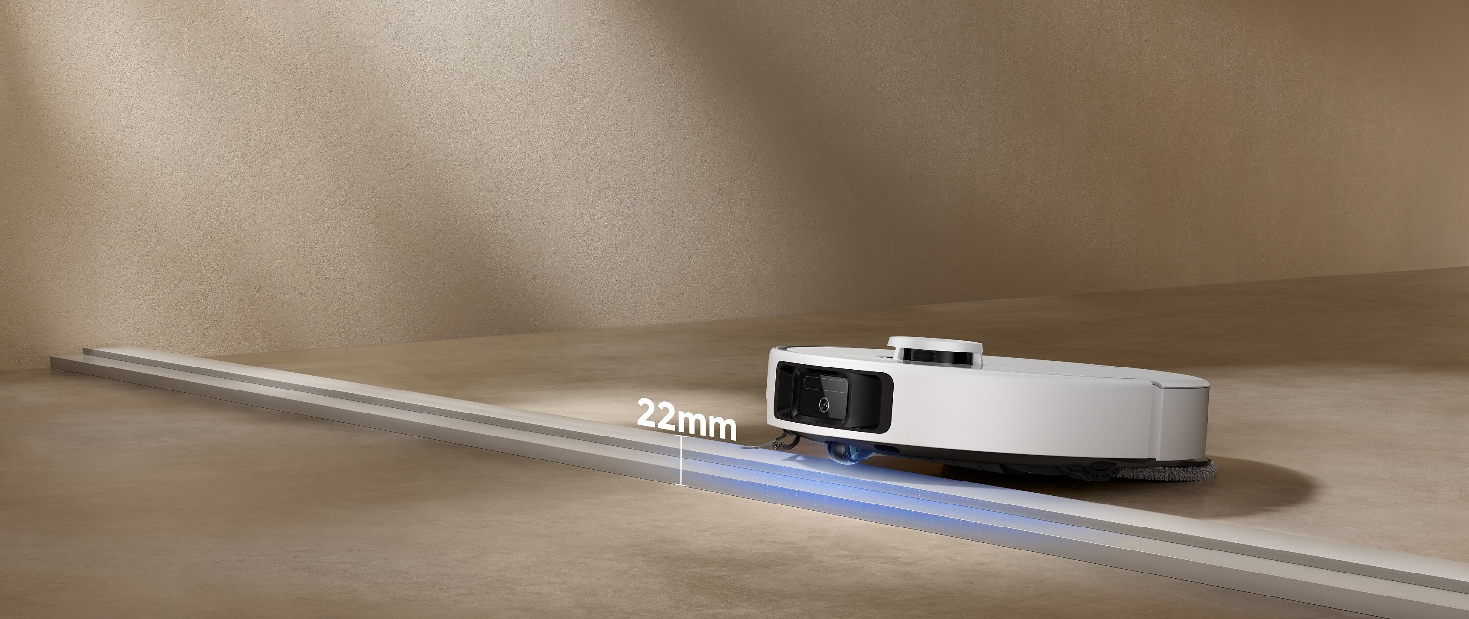 Biały robot odkurzający na jasnobrązowej podłodze. Jest na srebrnym torze. Znak wskazuje wysokość 22 mm.