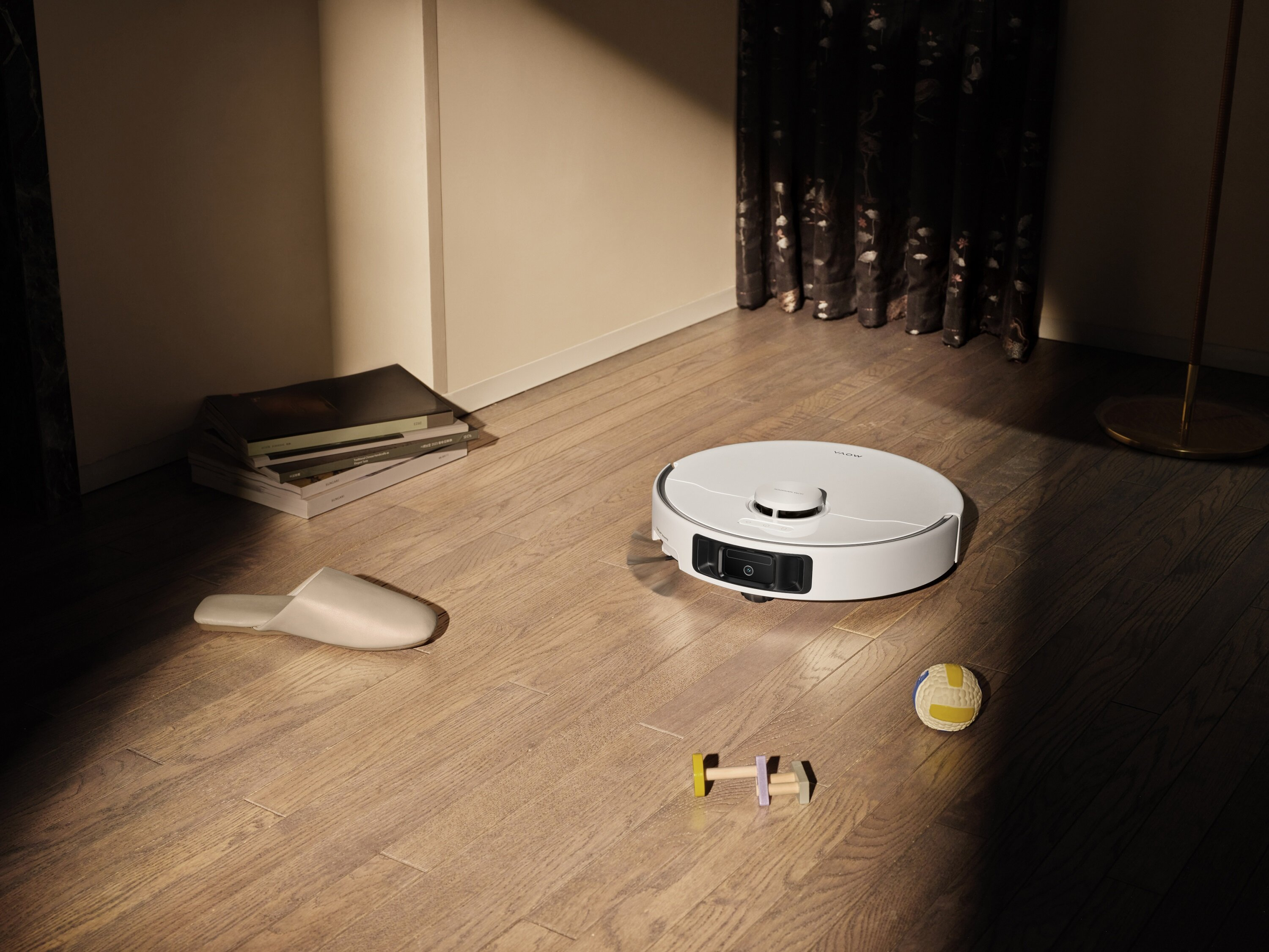 Biały robot odkurzający na drewnianej podłodze. Są też kapcie, zabawka, piłka i książki.
