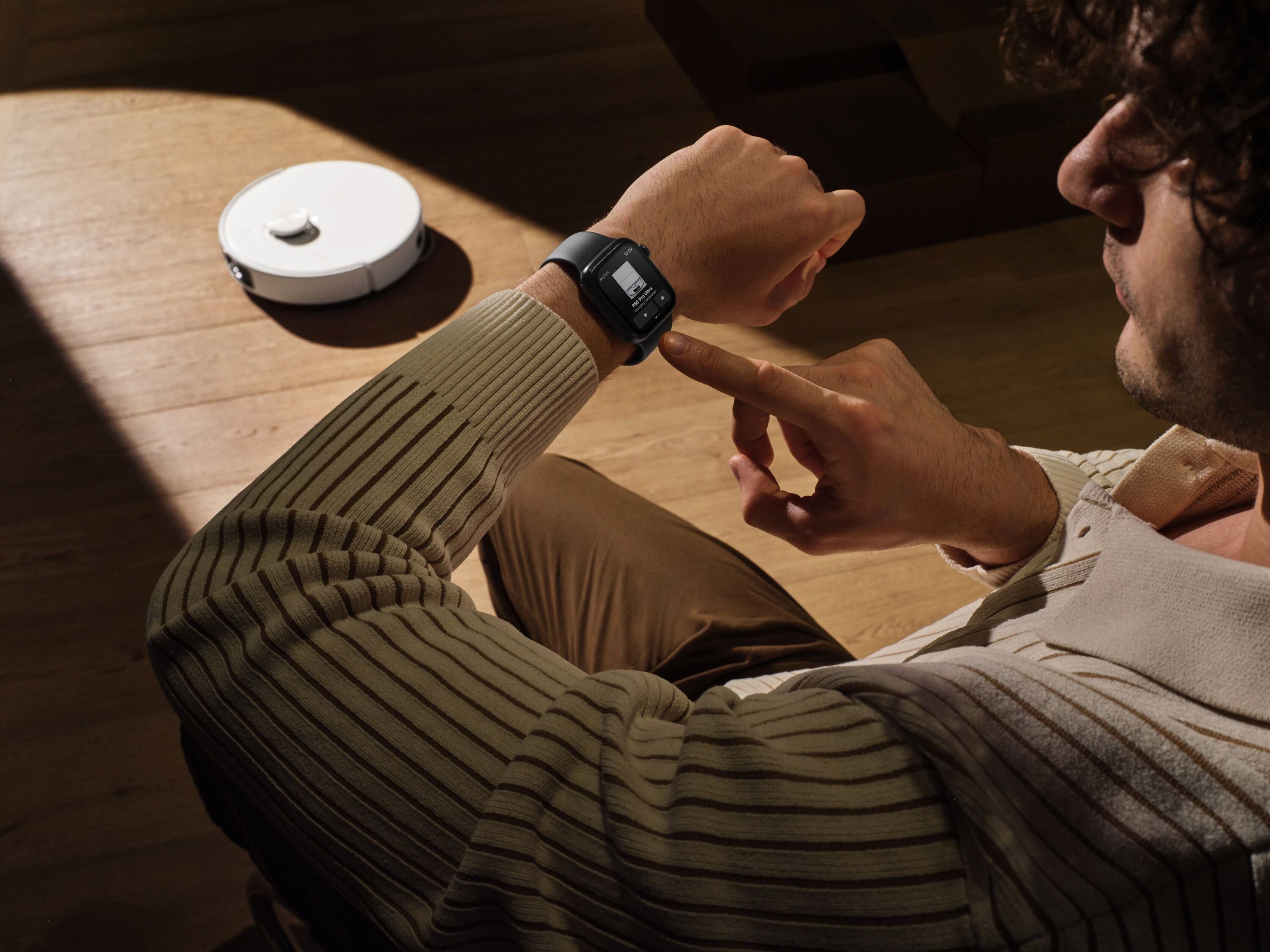 Osoba sprawdza smartwatch z aplikacją do sterowania robotem odkurzającym. Biały robot odkurzający znajduje się na drewnianej podłodze.