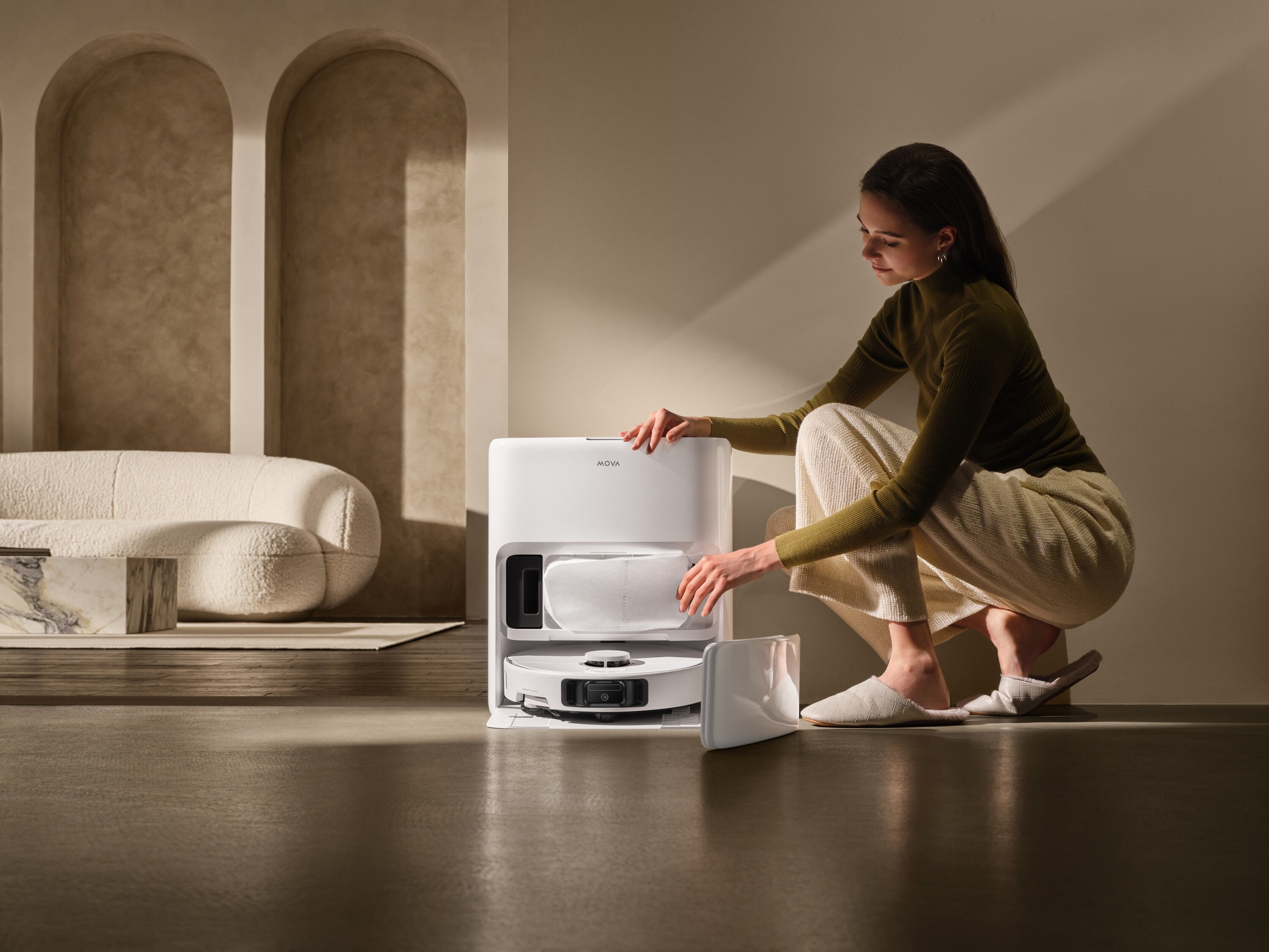 Kobieta kuca obok białego robota odkurzającego, otwierając komorę. Neutralny pokój z łukami i sofą.