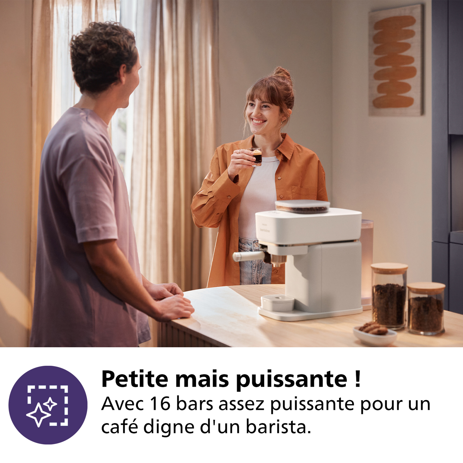 Een vrouw glimlacht, met koffie. Een man kijkt haar aan. Een wit koffiezetapparaat staat op een houten aanrecht. Koffiepotten zijn zichtbaar.