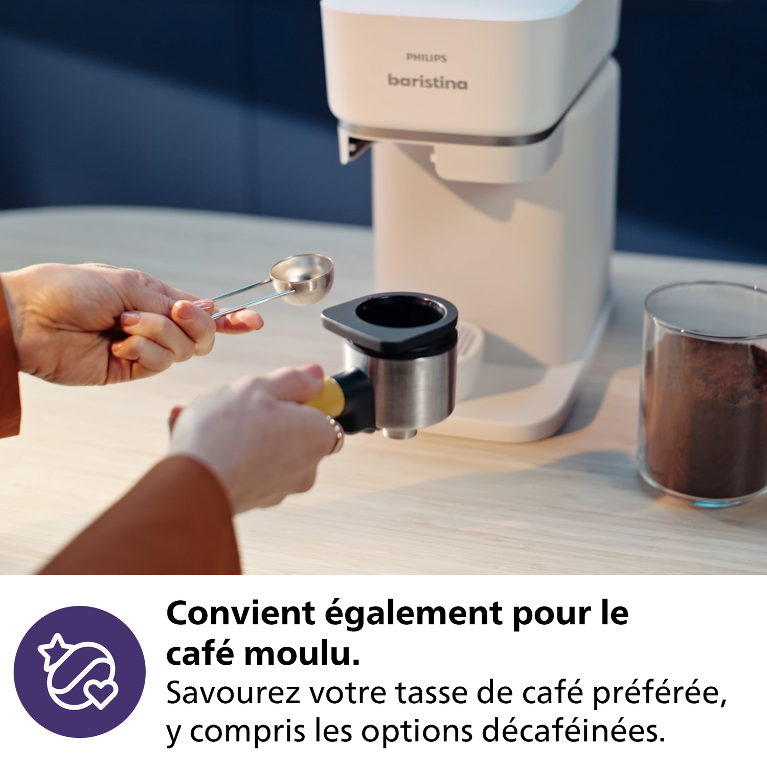 Een persoon gebruikt een koffiemachine. Een schepje wordt boven een filterhouder gehouden, een glas gemalen koffie staat op de achtergrond.