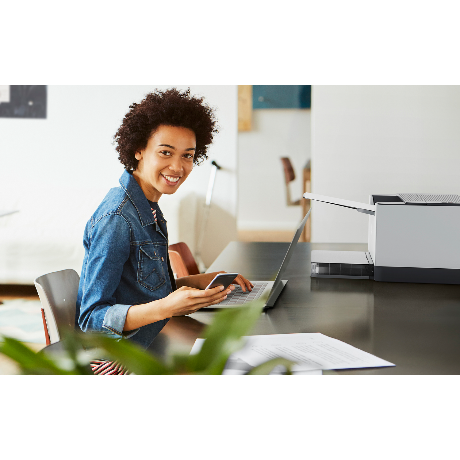 Woman with afro smiles at camera while working on laptop and phone. Printer and papers on the table.