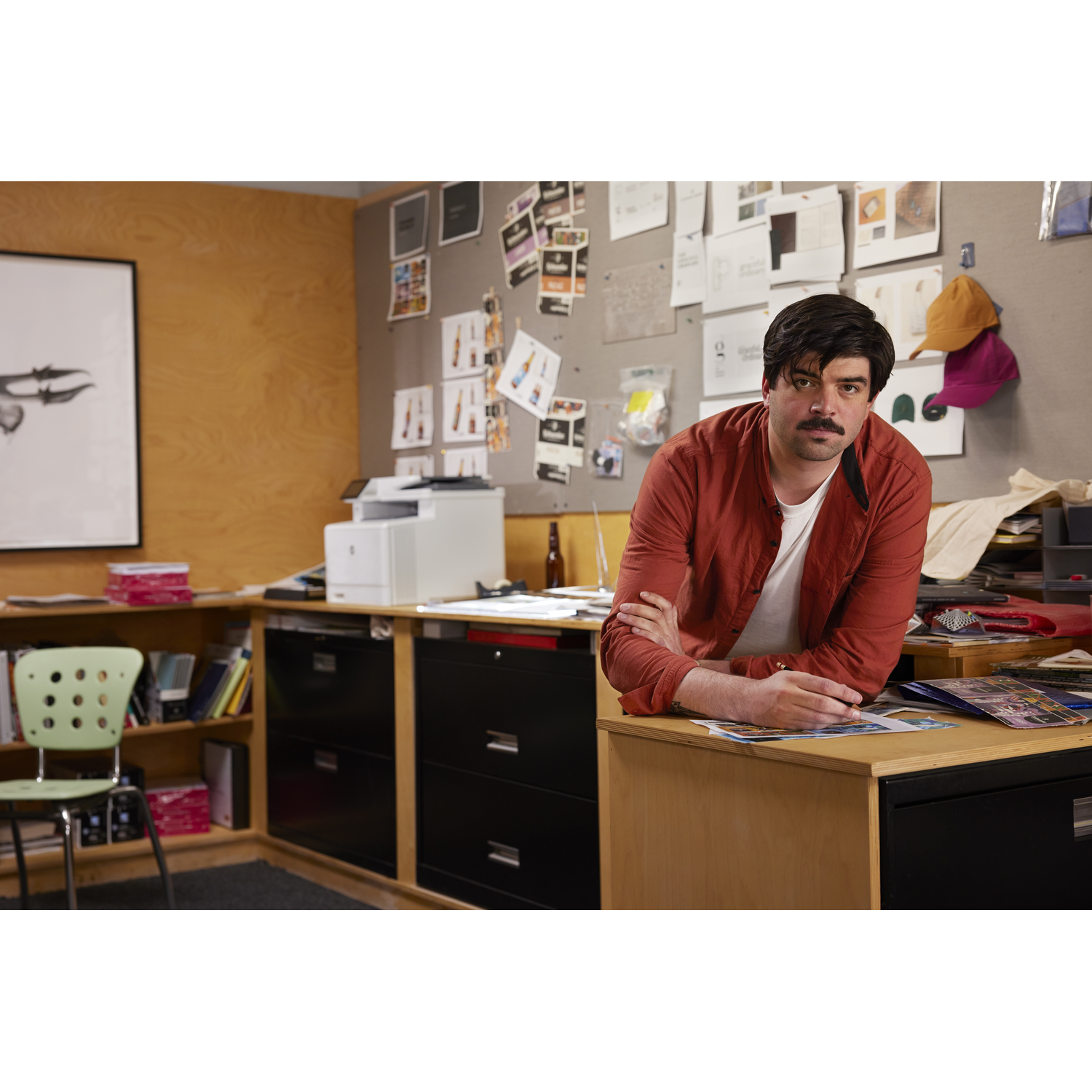 Man in orange shirt leans on desk, arms crossed. Office setting with papers, printer, and a green chair.