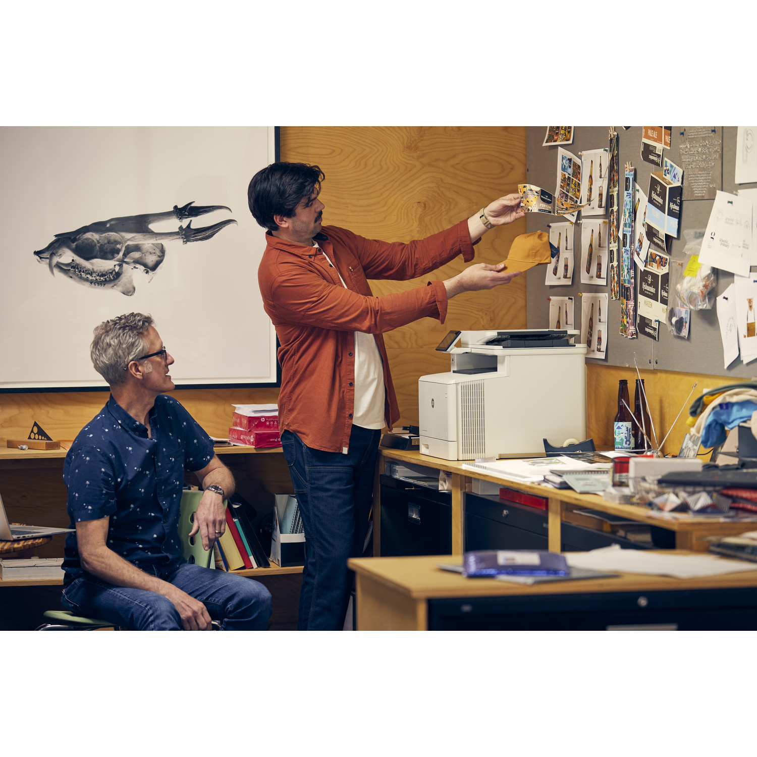 Two men in an office. One holds a cap, the other looks on. A printer sits on a desk, a skull art piece is behind them.