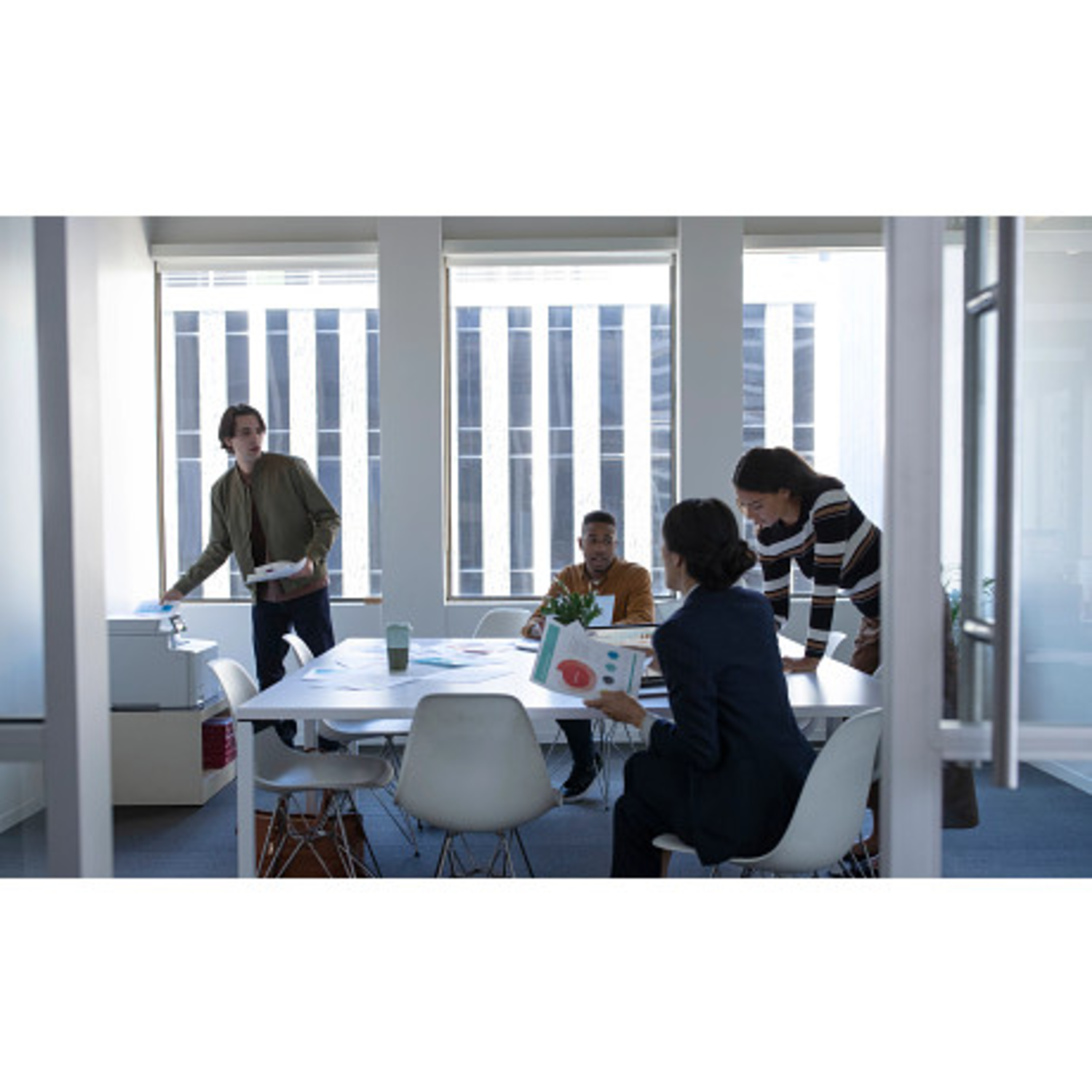 Four people in an office meeting. One man near printer, others seated around a table with papers. Windows in the background.