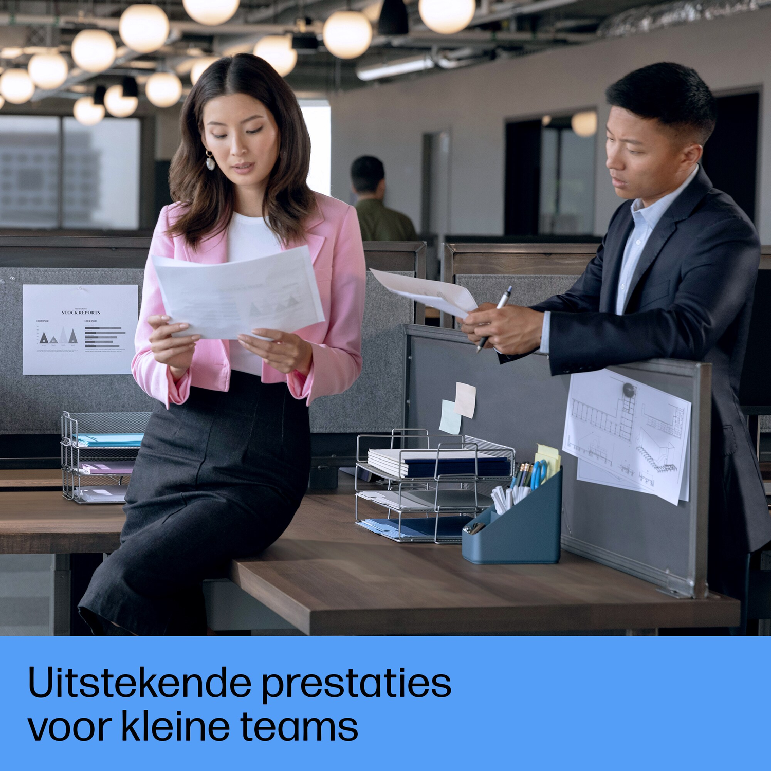 Twee mensen op kantoor bekijken documenten. Een vrouw in roze blazer zit, een man in pak staat. Kantooromgeving met bovenverlichting.
