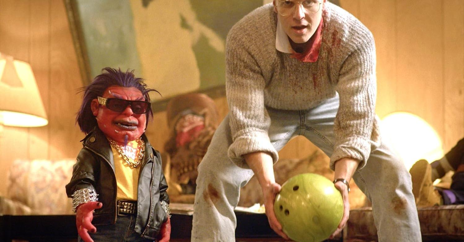 Ein Mann mit Blut auf seinem Pullover bereitet sich auf das Bowling vor. Eine Puppe mit Sonnenbrille und Lederjacke steht neben ihm.