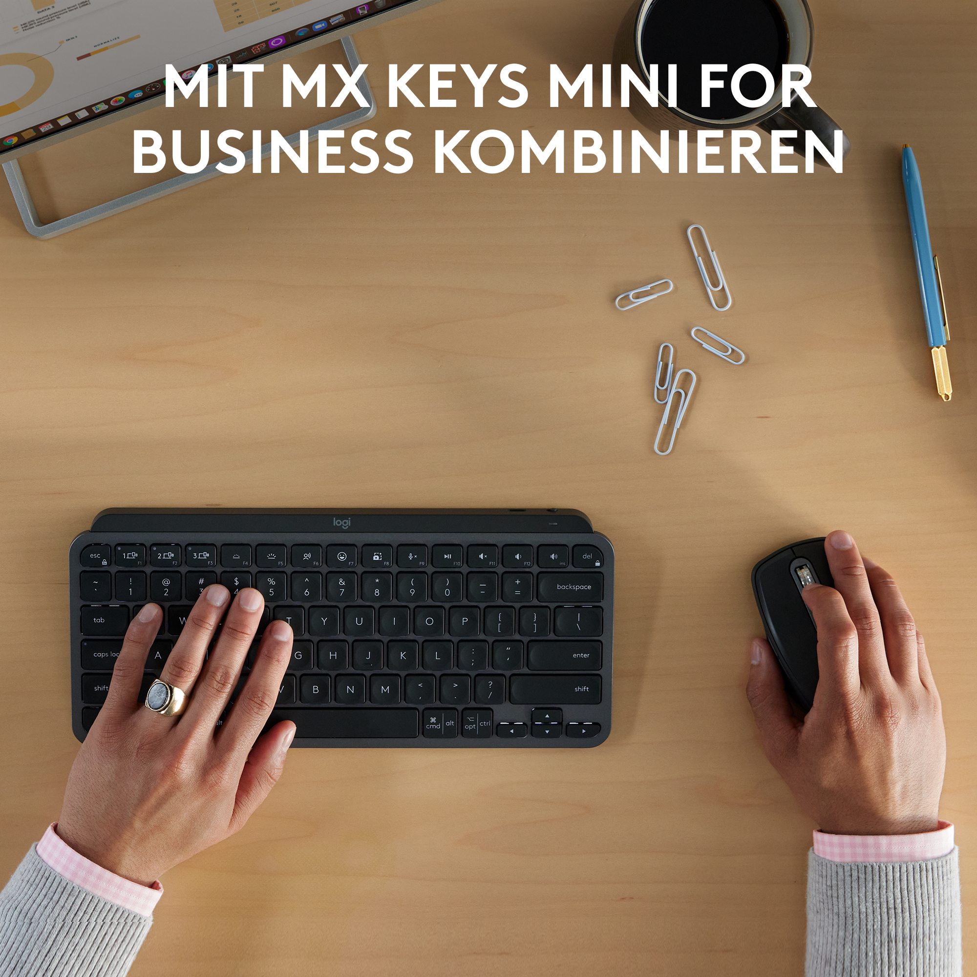 Ein Holztisch mit Tastatur, Maus, Stift und Kaffeetasse. Hände benutzen Tastatur und Maus. Büroklammern sind verstreut.
