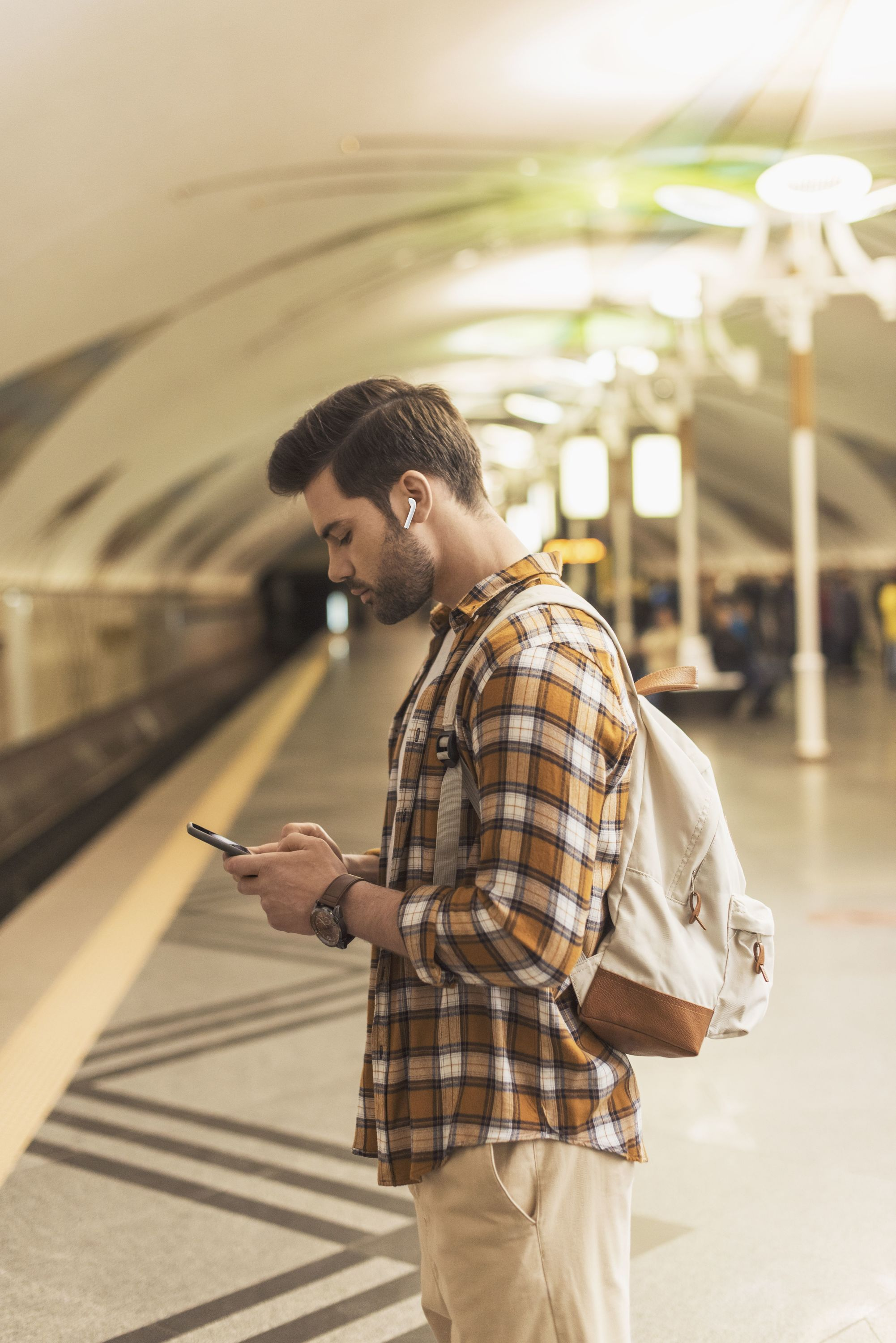 Mann im karierten Hemd in der U-Bahn, benutzt Telefon, mit Rucksack. Hintergrund zeigt Bahnsteig, Lichter, andere Leute.
