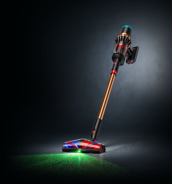 Odkurzacz bezprzewodowy z brązową rurą, czarnymi i czerwonymi detalami oraz niebieskim wierzchem. Zielone światło oświetla podłogę.