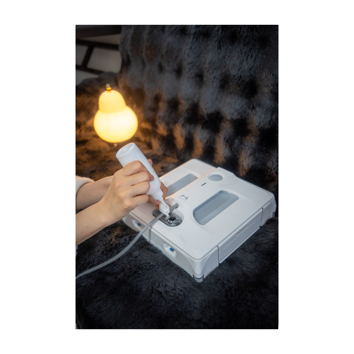 Mani che versano liquido da una bottiglia bianca in un robot aspirapolvere bianco su una superficie nera e soffice, con una lampada gialla sullo sfondo.