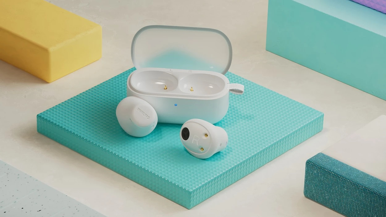 Białe bezprzewodowe słuchawki z etui ładującym na turkusowej powierzchni. Żółte i niebieskie bloki w tle. Słuchawki są oznakowane.