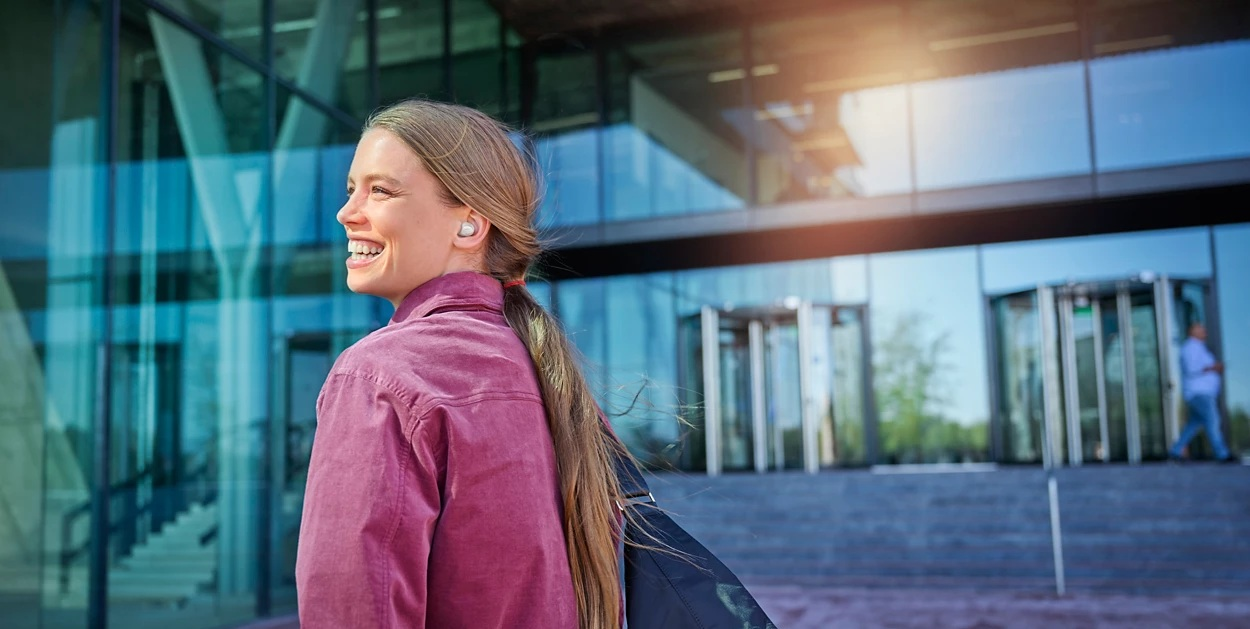 Uśmiechnięta kobieta ze słuchawkami przechodzi obok nowoczesnego szklanego budynku, niosąc torbę. W tle widać drzwi obrotowe.