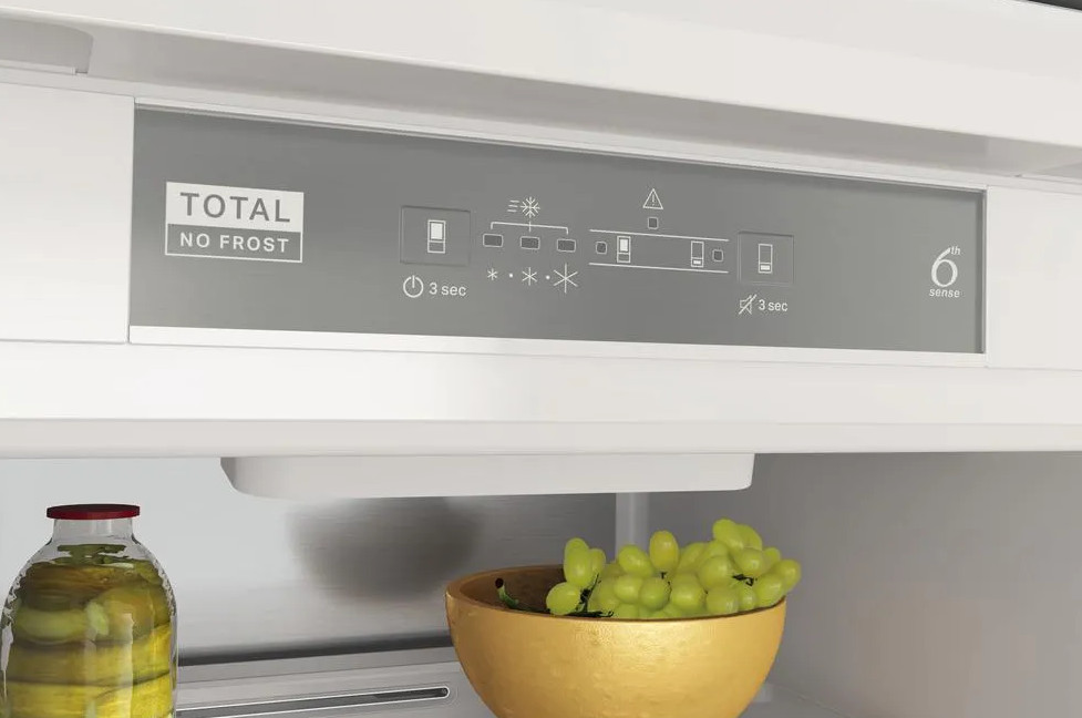 Wewnątrz lodówki: panel sterowania z przyciskami, słoik, złota miska z winogronami. Białe wnętrze.