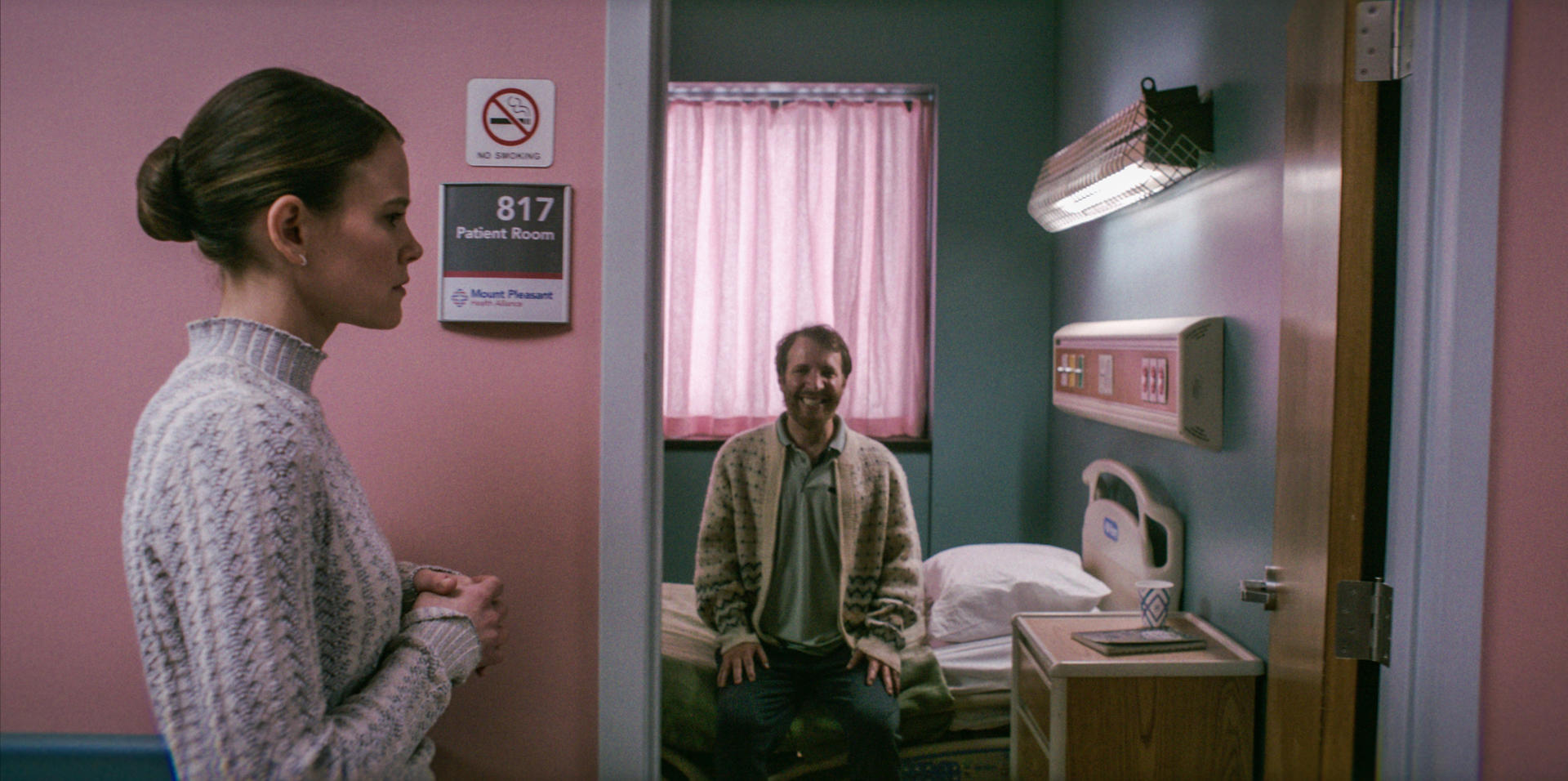 Eine Frau steht an einer Patientenzimmertür. Ein Mann sitzt drinnen. Das Zimmer hat ein Bett, ein Fenster mit rosa Vorhängen und medizinische Geräte.