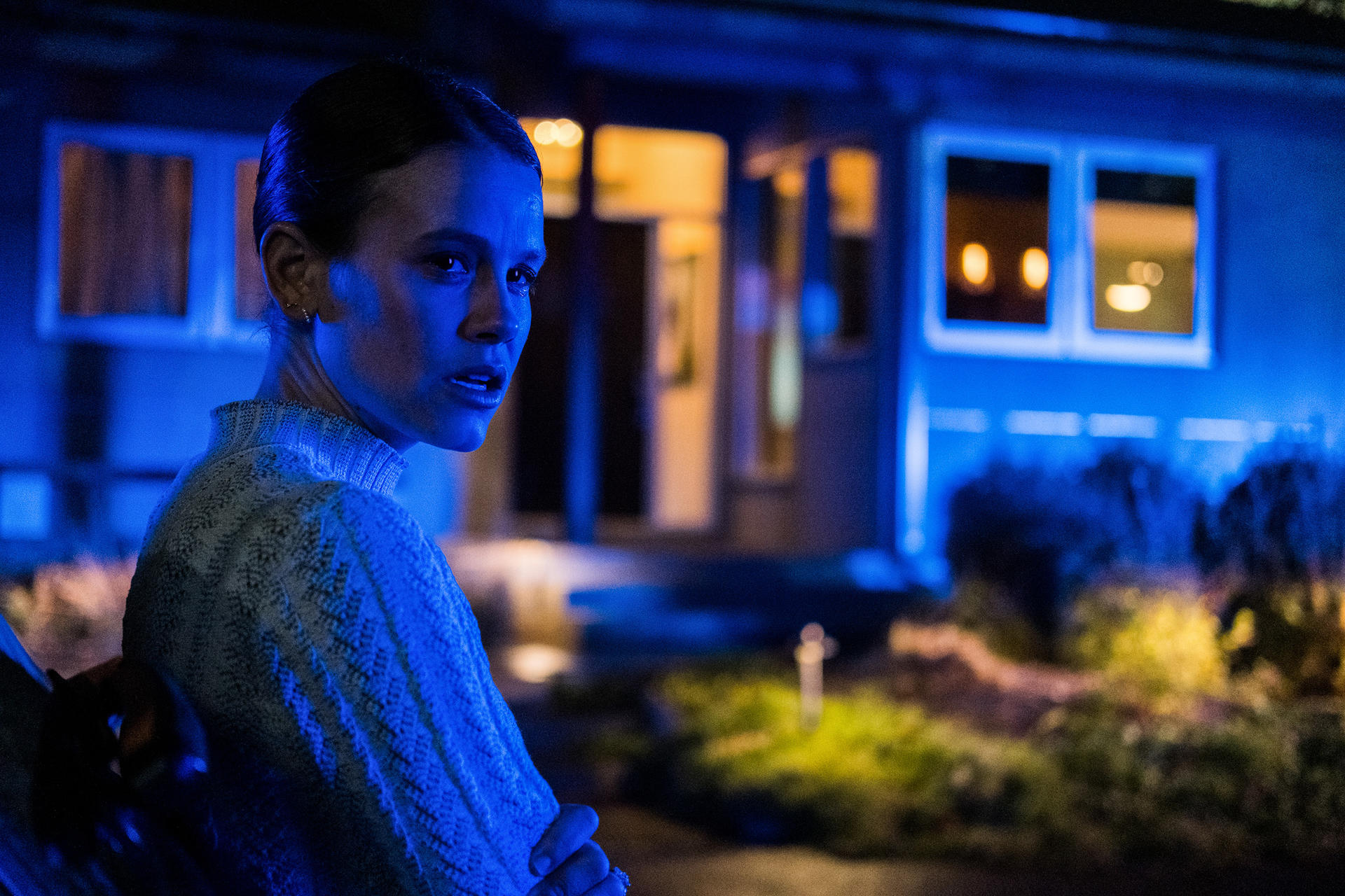 Eine Frau in einer blau beleuchteten Szene, die besorgt aussieht. Sie trägt einen hellen Pullover. Ein Haus mit beleuchteten Fenstern ist im Hintergrund.