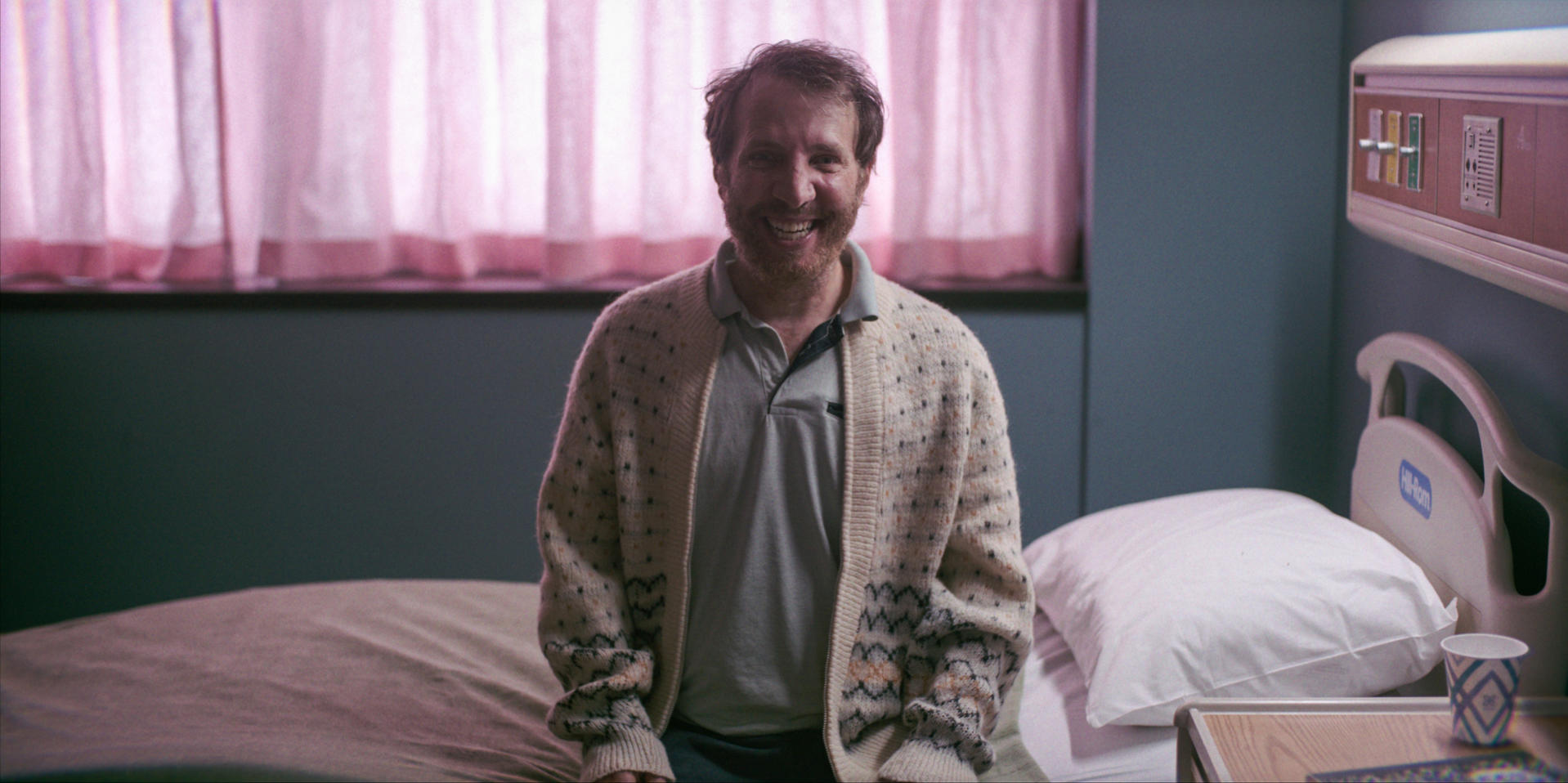 Mann in einem Krankenhauszimmer, lächelnd, mit einem gemusterten Pullover. Pinke Vorhänge im Hintergrund und ein Krankenhausbett.