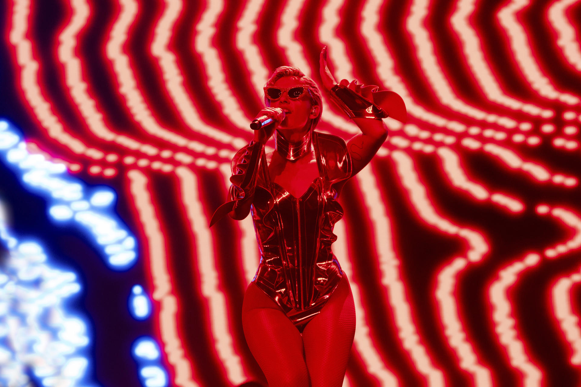 Eine Darstellerin in einem roten Outfit singt in ein Mikrofon. Der Hintergrund hat rote und weiße Wellenlinien.