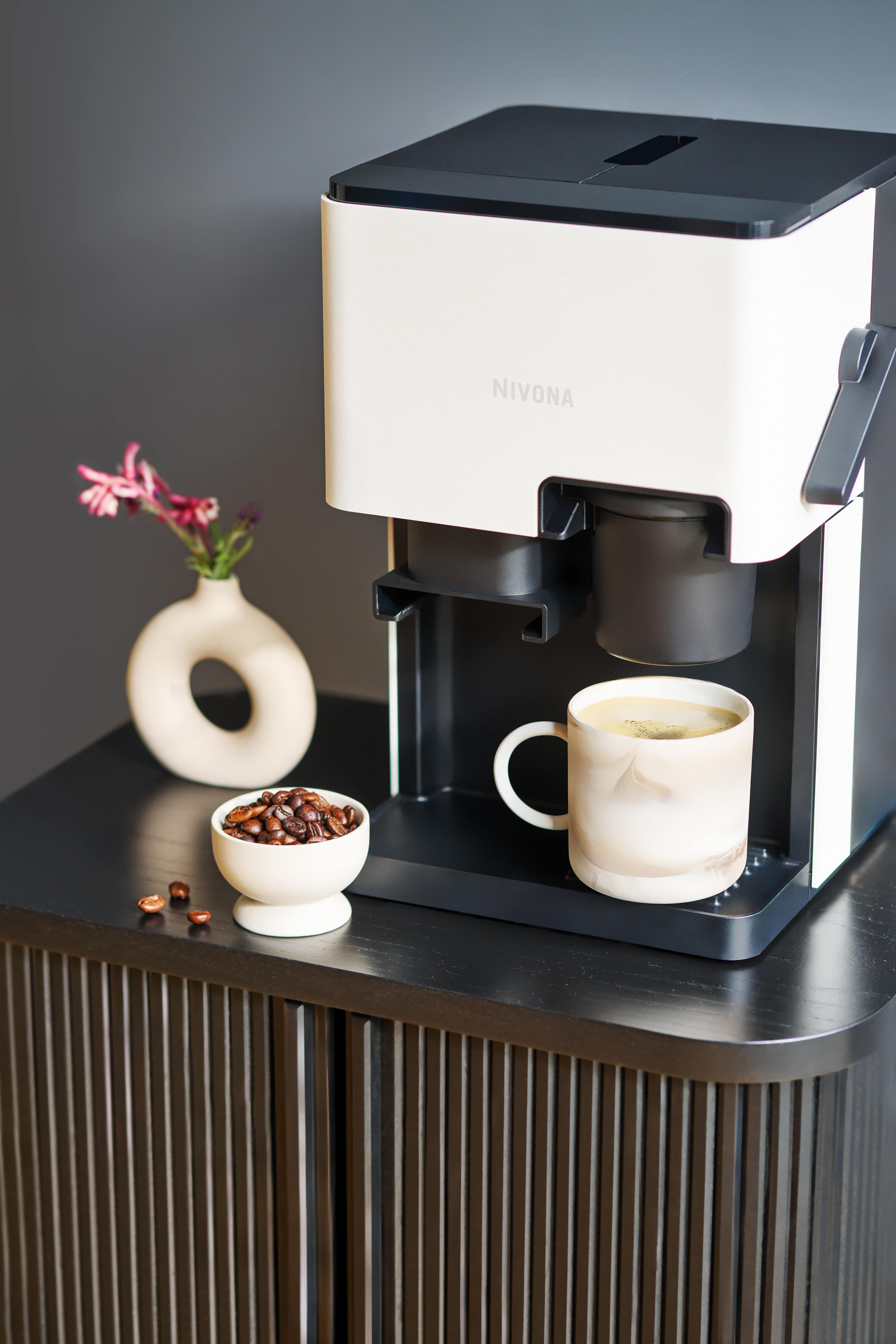 Ekspres do kawy z filiżanką kawy, miską ziaren kawy i wazonem z kwiatami, wszystko na drewnianej szafce.