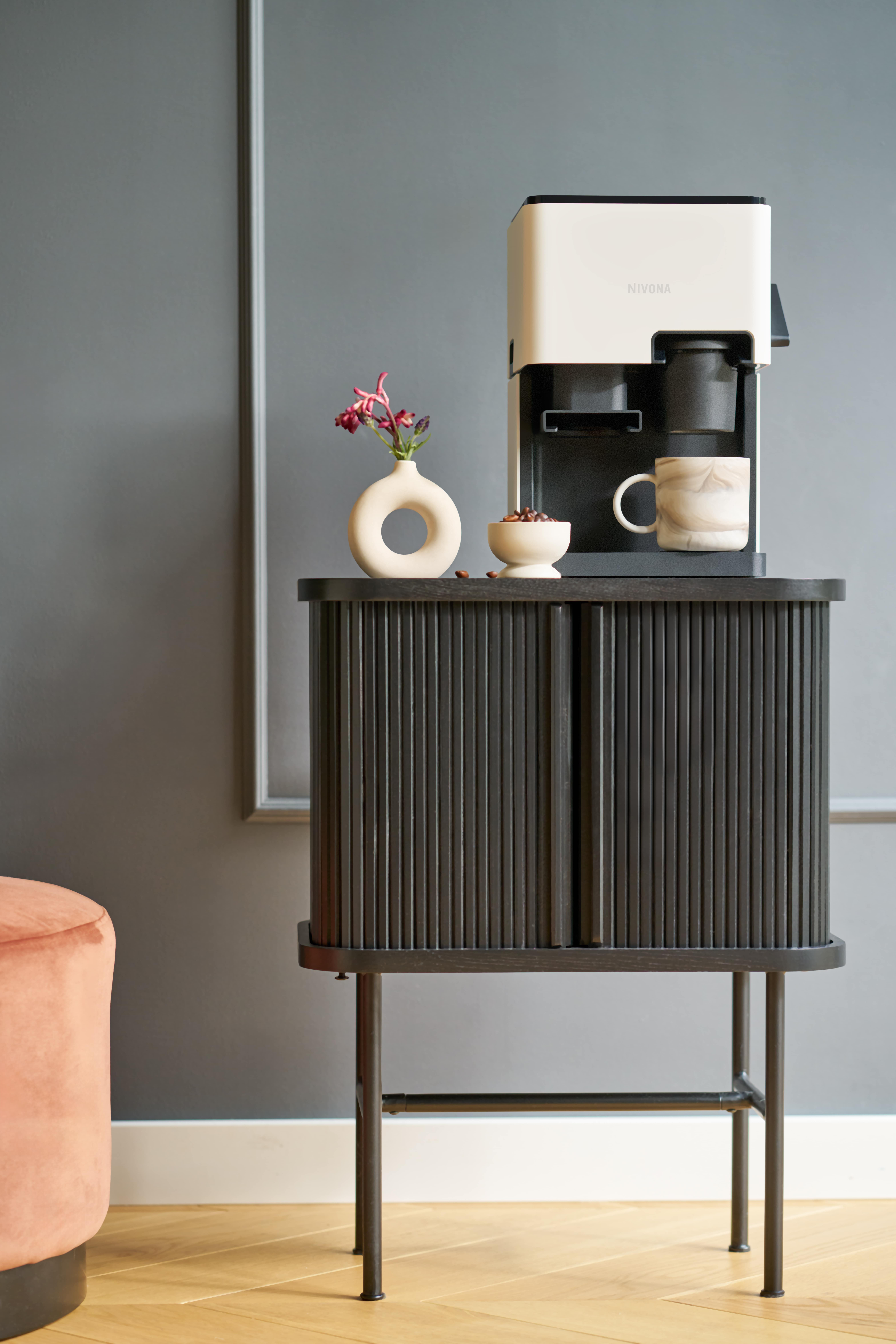 Ekspres do kawy na ciemnej szafce z wazonem z kwiatami, kubkiem i ziarnami. Różowy puf po lewej.