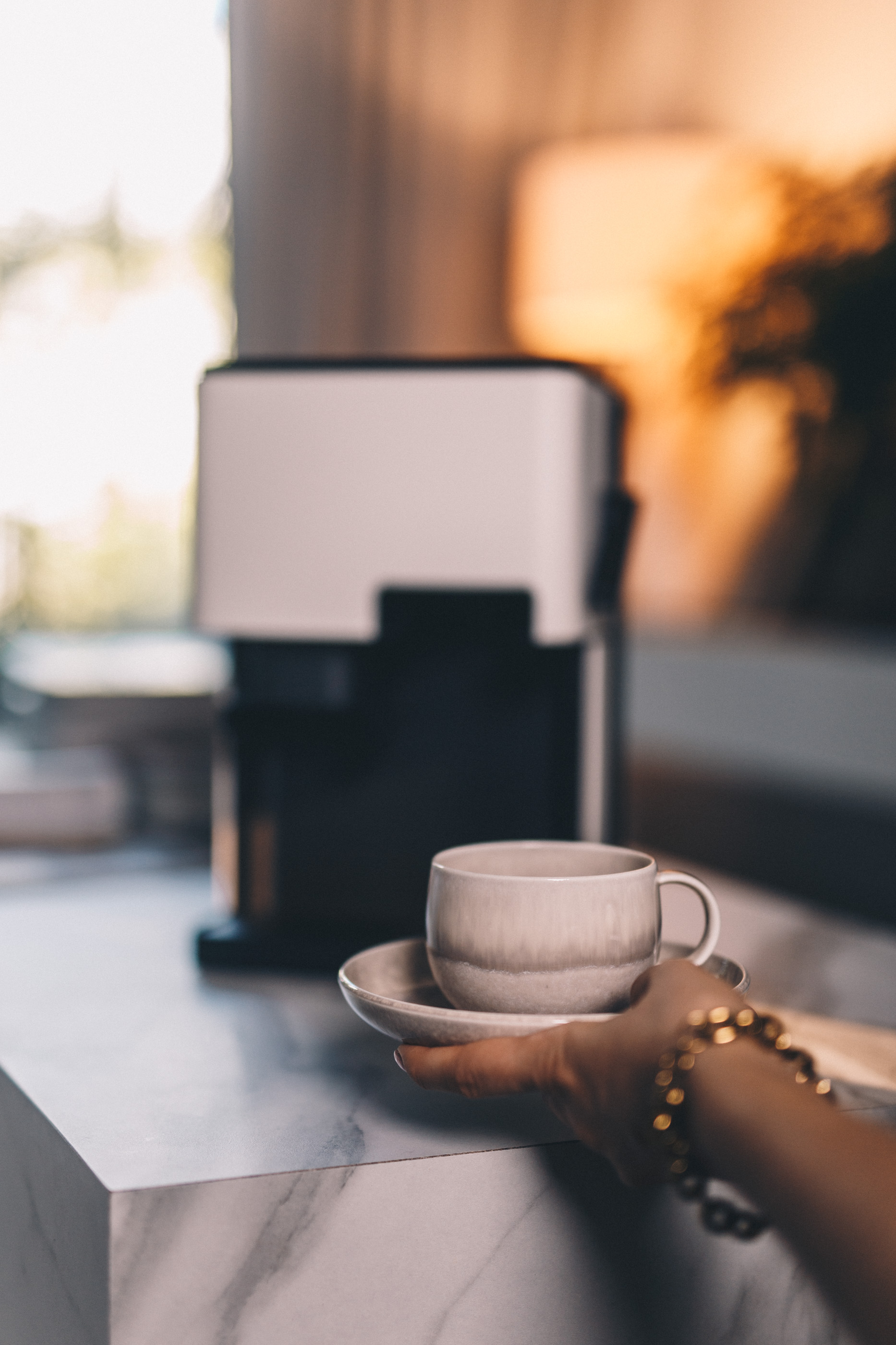 Dłoń trzymająca filiżankę ze spodkiem. Ekspres do kawy w tle. Złota bransoletka na nadgarstku.