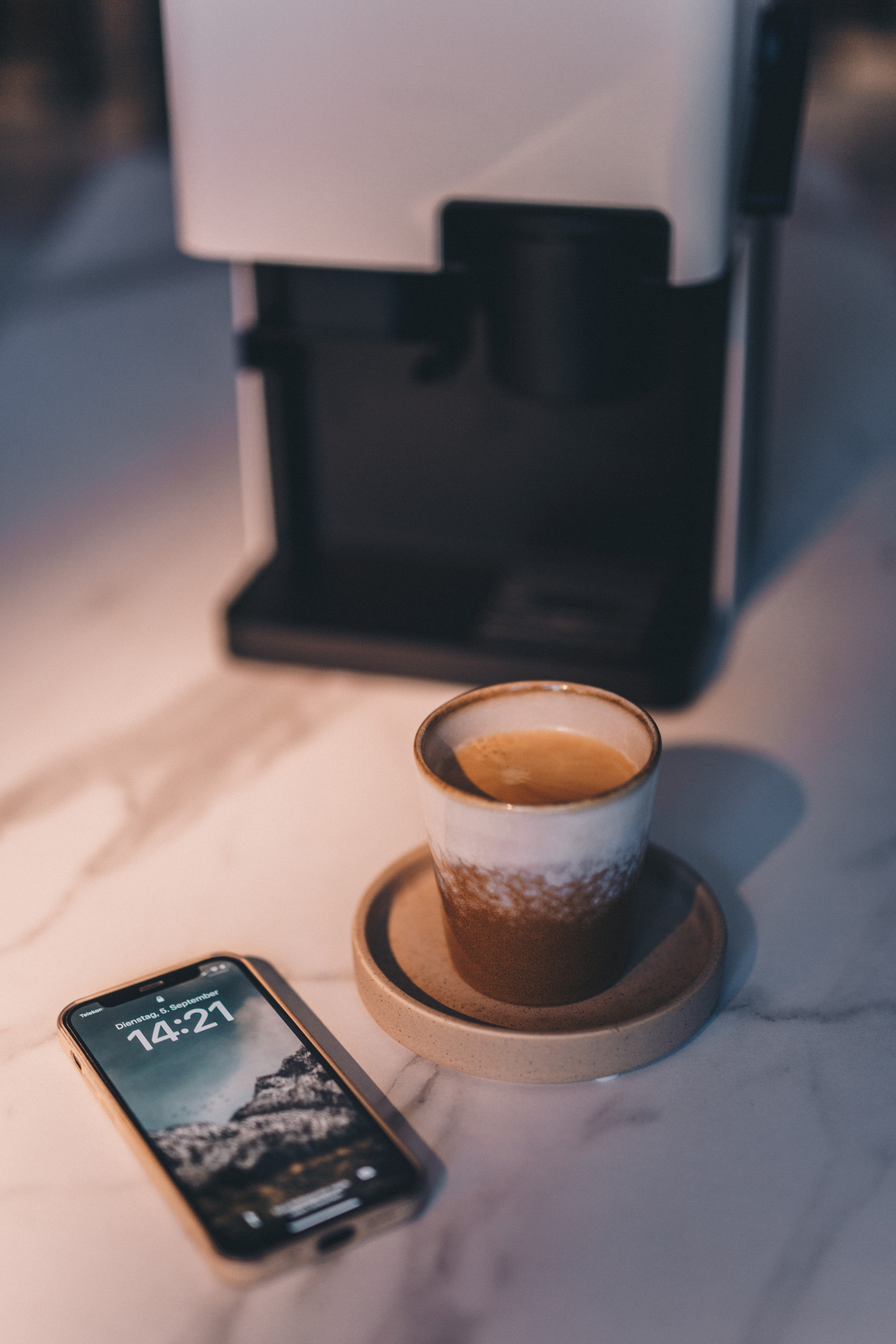 Filiżanka kawy na spodku, telefon z godziną i ekspres do kawy na marmurowej powierzchni.