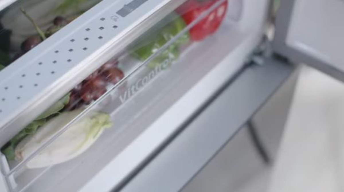 Szuflada lodówki wypełniona produktami: czerwony pomidor, zielona sałata, winogrona i endywia. Szuflada jest częściowo otwarta.