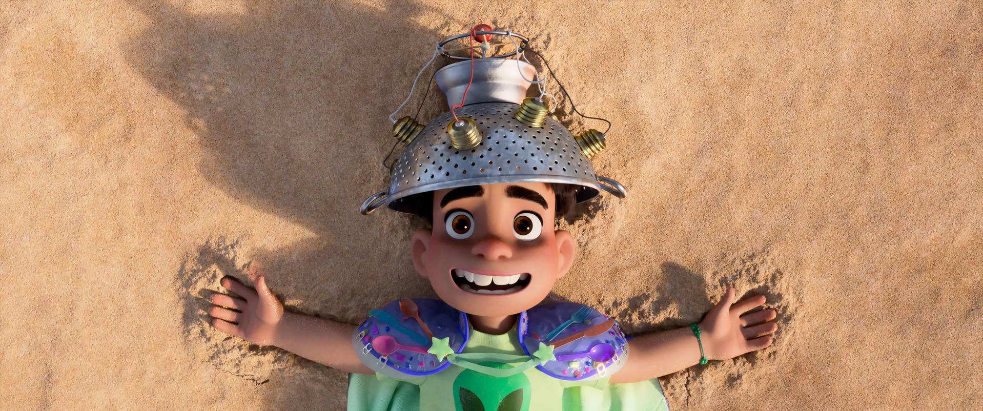 Ein lächelnder Junge liegt auf Sand und trägt einen Nudelsiebhelm. Er hat ein grünes Alien-Shirt und ein Armband. Der Hintergrund ist sandig.
