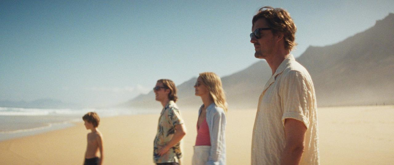 Eine Familie steht am Strand. Der Himmel ist blau. Im Hintergrund sind Berge. Ein Kind und drei Erwachsene stehen im Profil.