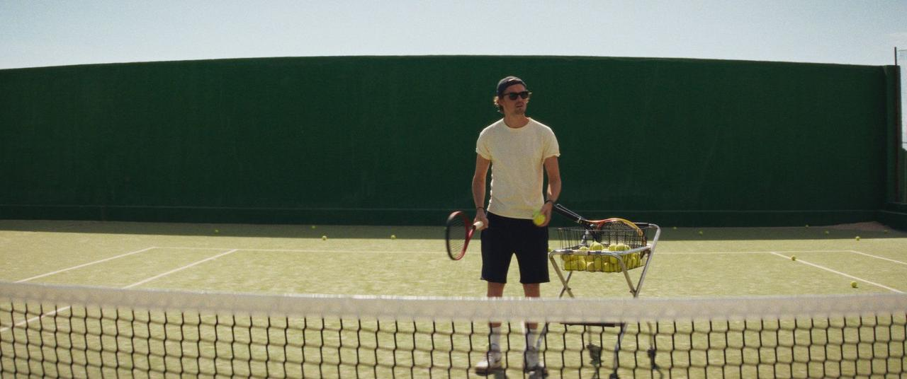Ein Mann steht auf einem Tennisplatz und hält einen Tennisschläger und einen Ball. Ein Wagen mit Tennisbällen und ein Netz sind sichtbar.