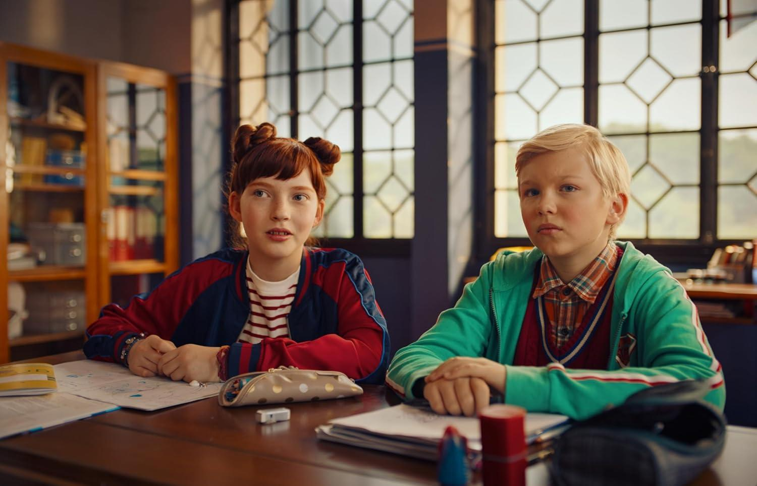 Zwei Kinder sitzen in einem Klassenzimmer an einem Schreibtisch. Ein Mädchen mit roten Haaren und ein Junge mit blonden Haaren schauen nach vorne. Bücherregale sind sichtbar.