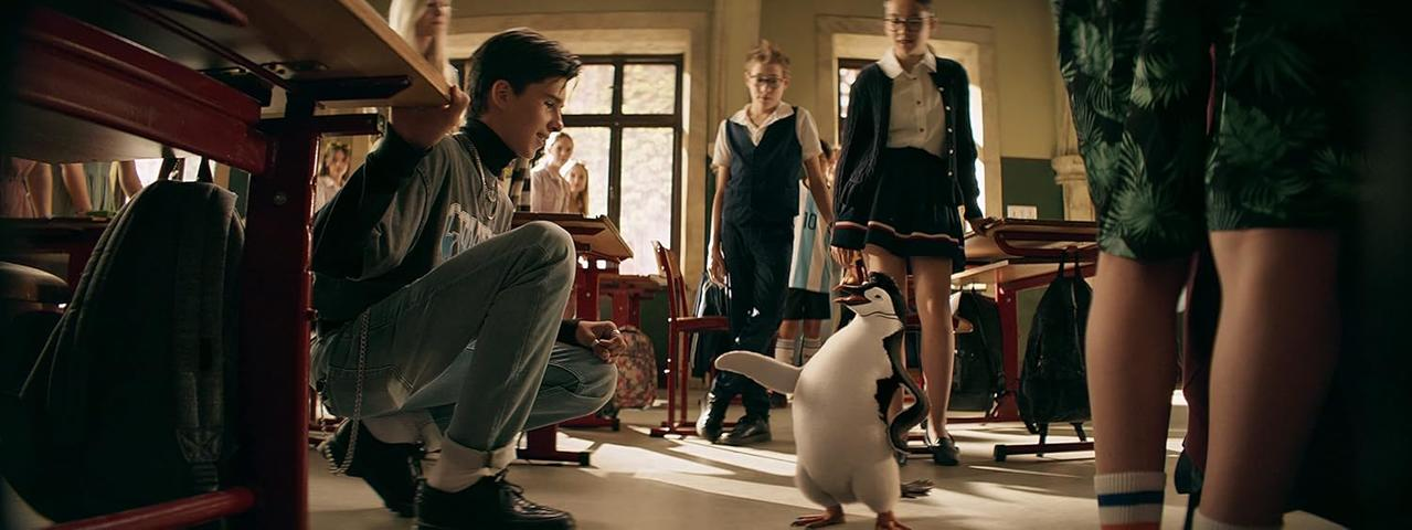 Ein Schüler kauert und betrachtet einen Pinguin, der in einem Klassenzimmer steht. Andere Schüler und Tische sind sichtbar.