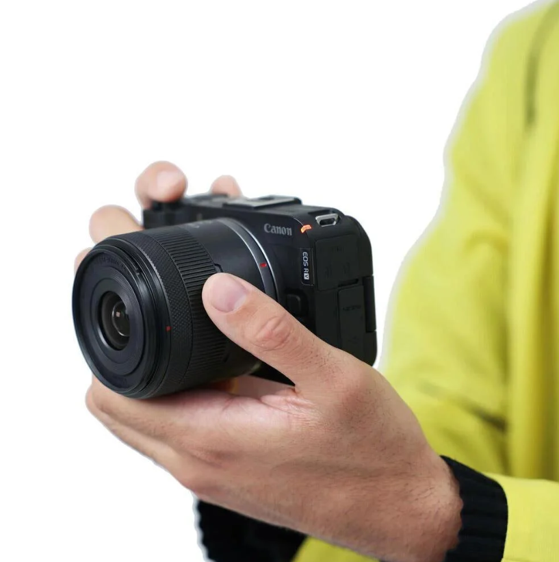 Bir kişi siyah bir Canon EOS R kamera tutuyor. Kişi sarı bir gömlek giyiyor. Kamera lensi izleyiciye dönük.