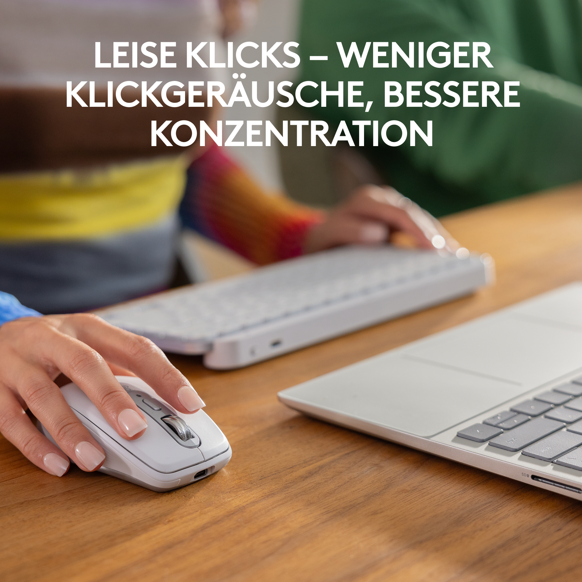 Eine Hand benutzt eine weiße Maus. Eine Tastatur und ein Laptop befinden sich im Hintergrund auf einem Holztisch.