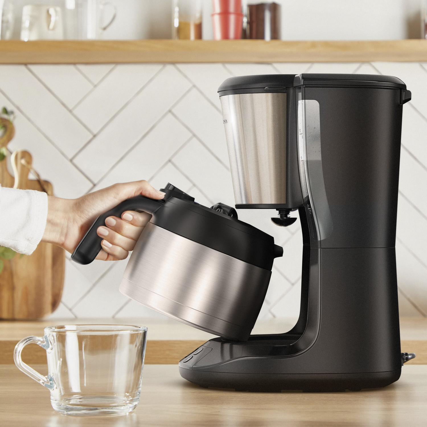 A person is holding a coffee pot, pouring coffee into a glass mug. The coffee machine is black and silver on a wooden surface.