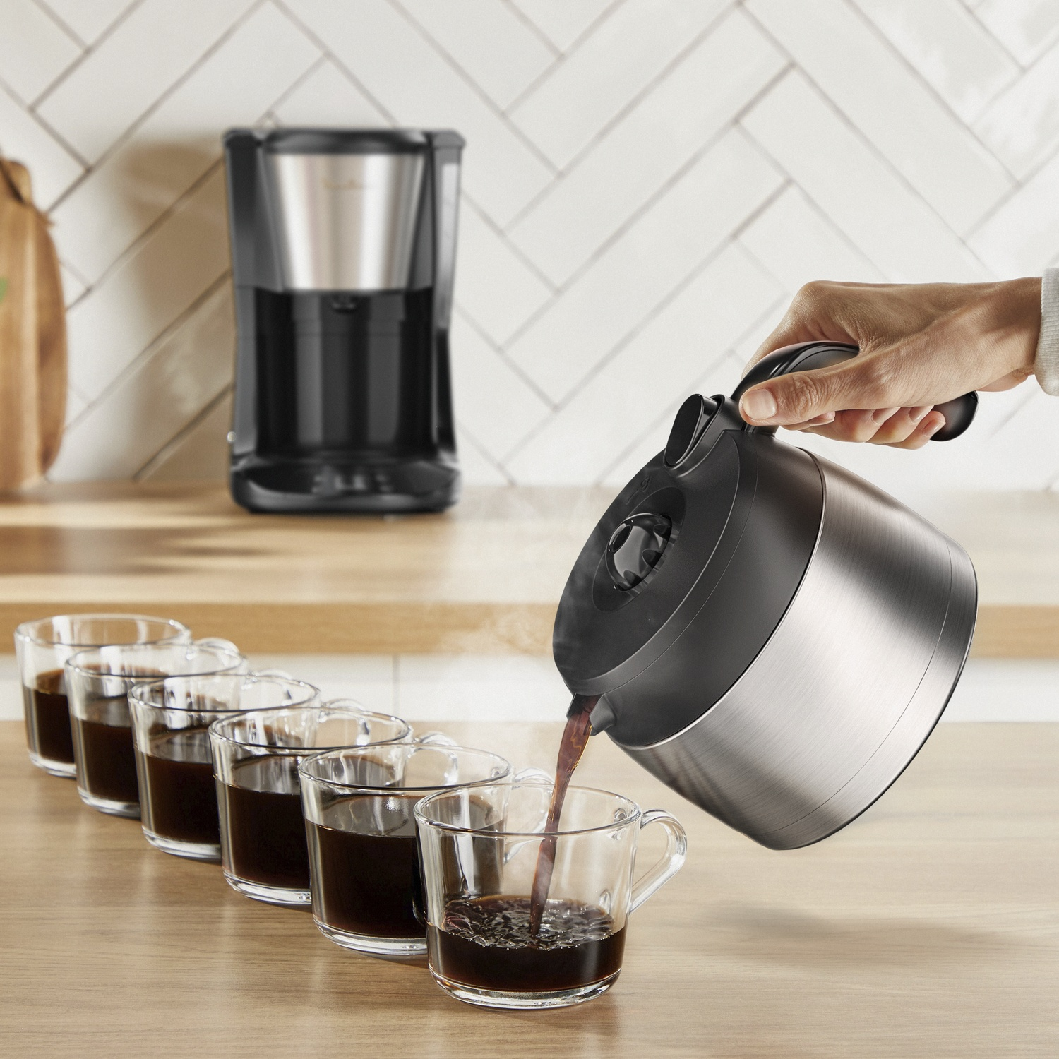 A person pours coffee from a silver carafe into a clear glass. Six glasses of coffee sit in a row. A coffee maker is in the background.