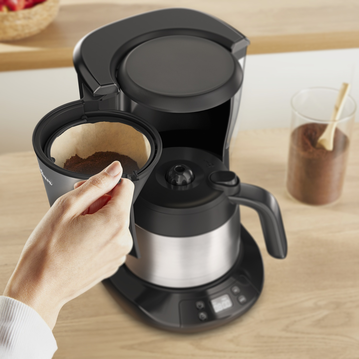 A person is placing a filter into a black coffee maker. Ground coffee fills the filter. A glass jar of coffee is in the background.