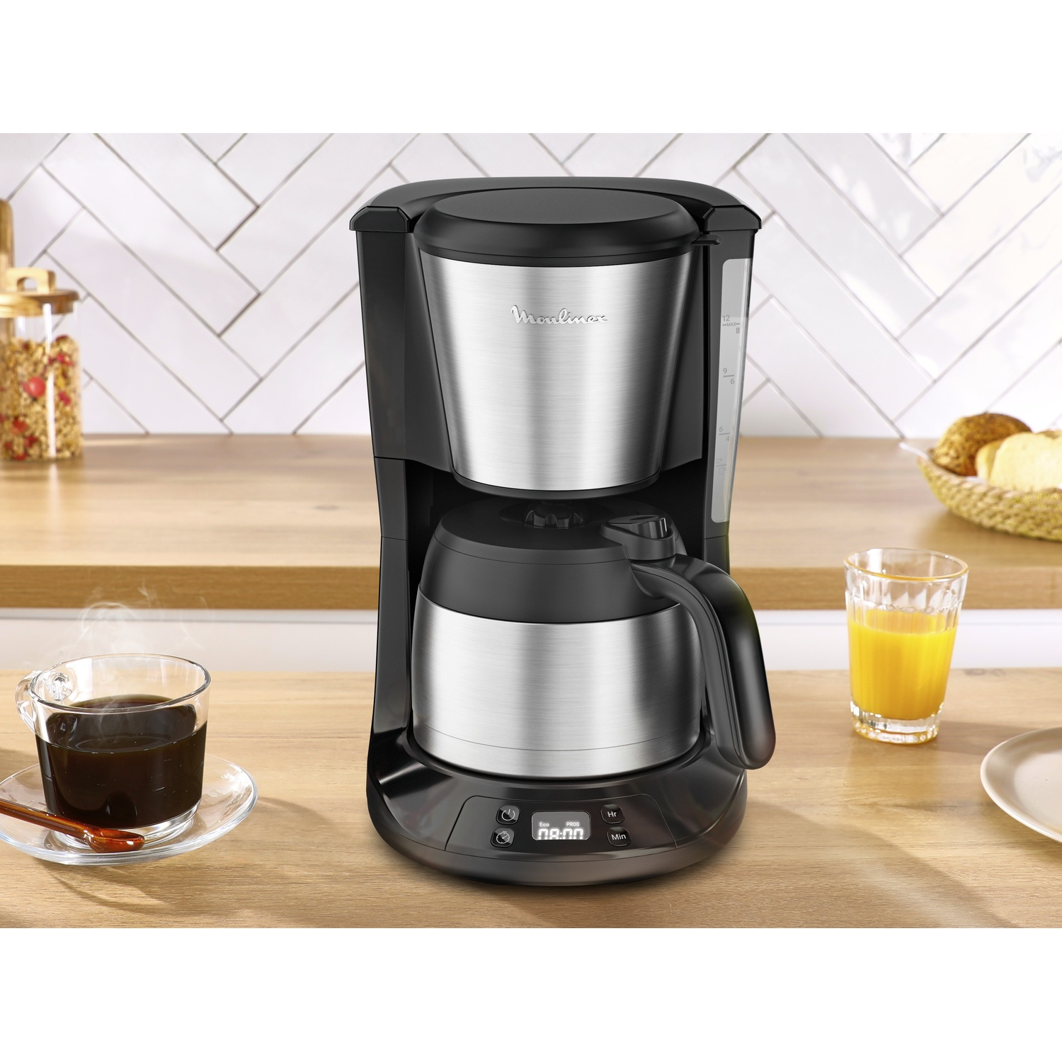 Coffee maker on a wooden surface with a cup of coffee, a glass of juice, and bread in a basket. White tile background.