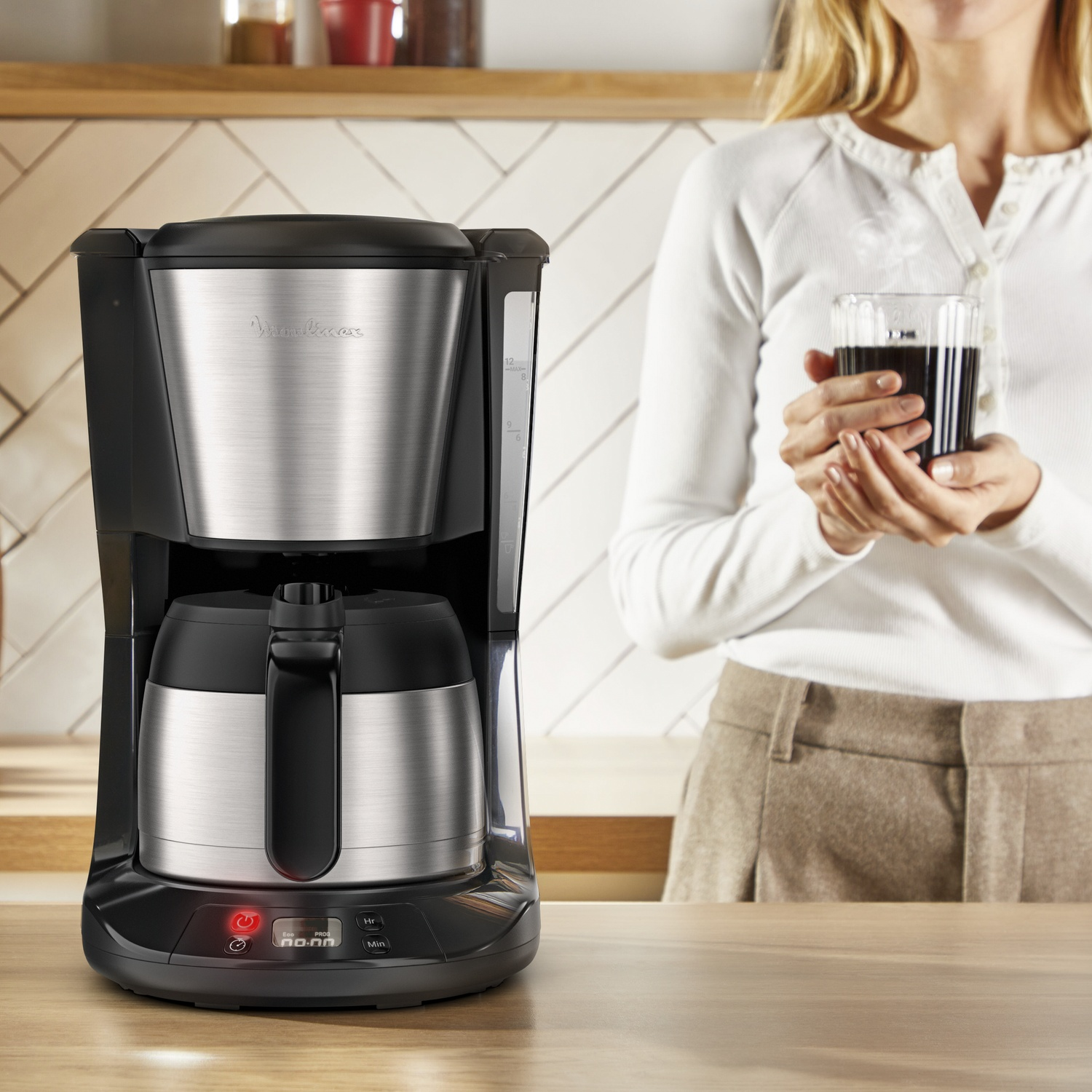 A coffee maker with a woman holding a coffee. The coffee maker is black and silver. The woman wears a white shirt and brown pants.