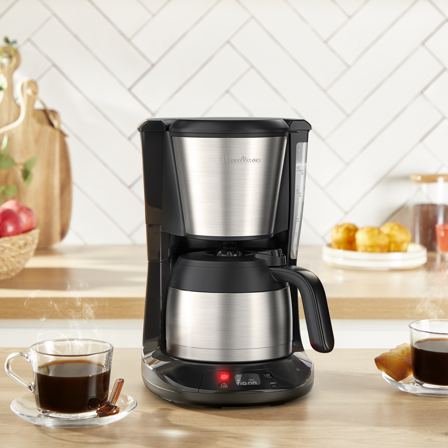 A coffee maker with a glass cup of coffee on a wooden counter. Muffins, a cutting board, and a basket are also visible.