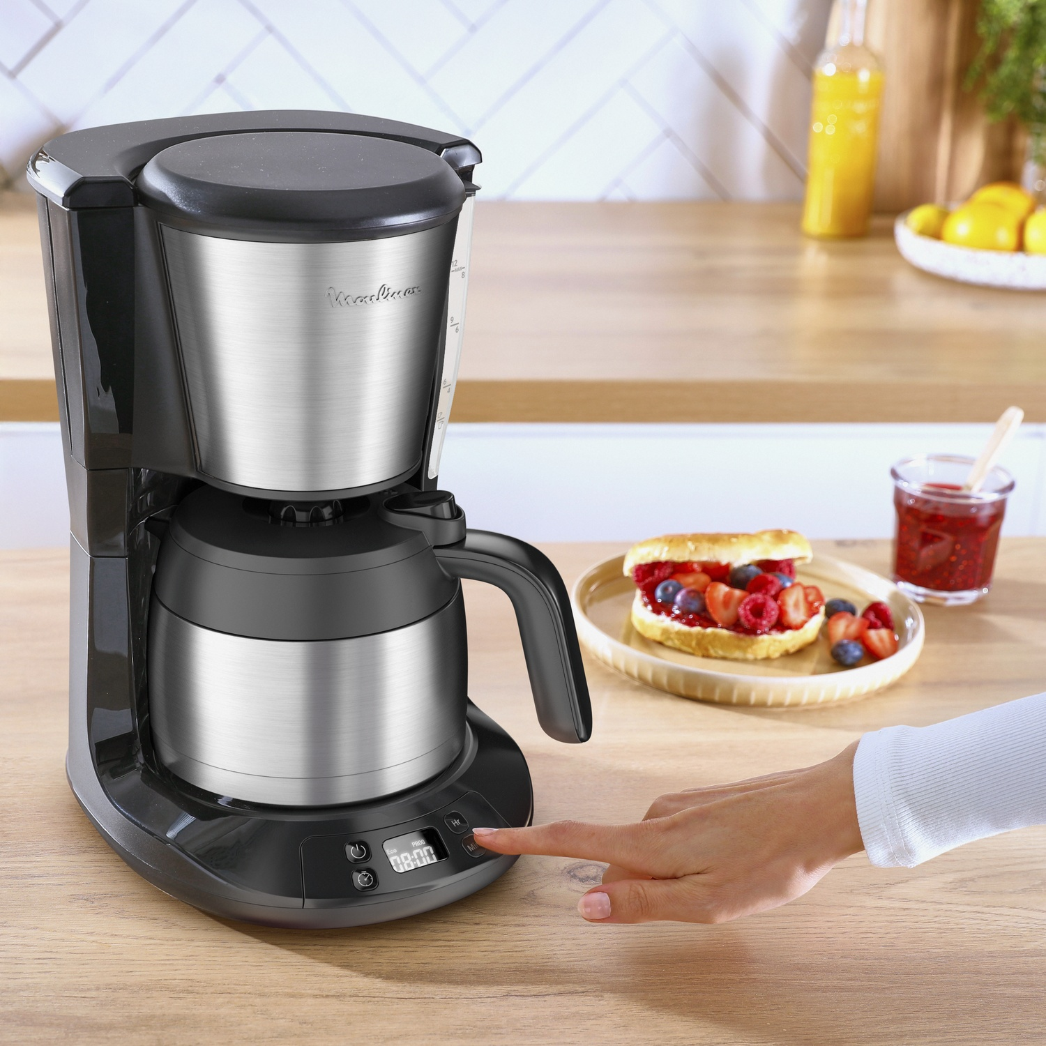 A black and silver coffee maker is on a wooden counter. A hand is pressing the button. A sandwich with berries is nearby.