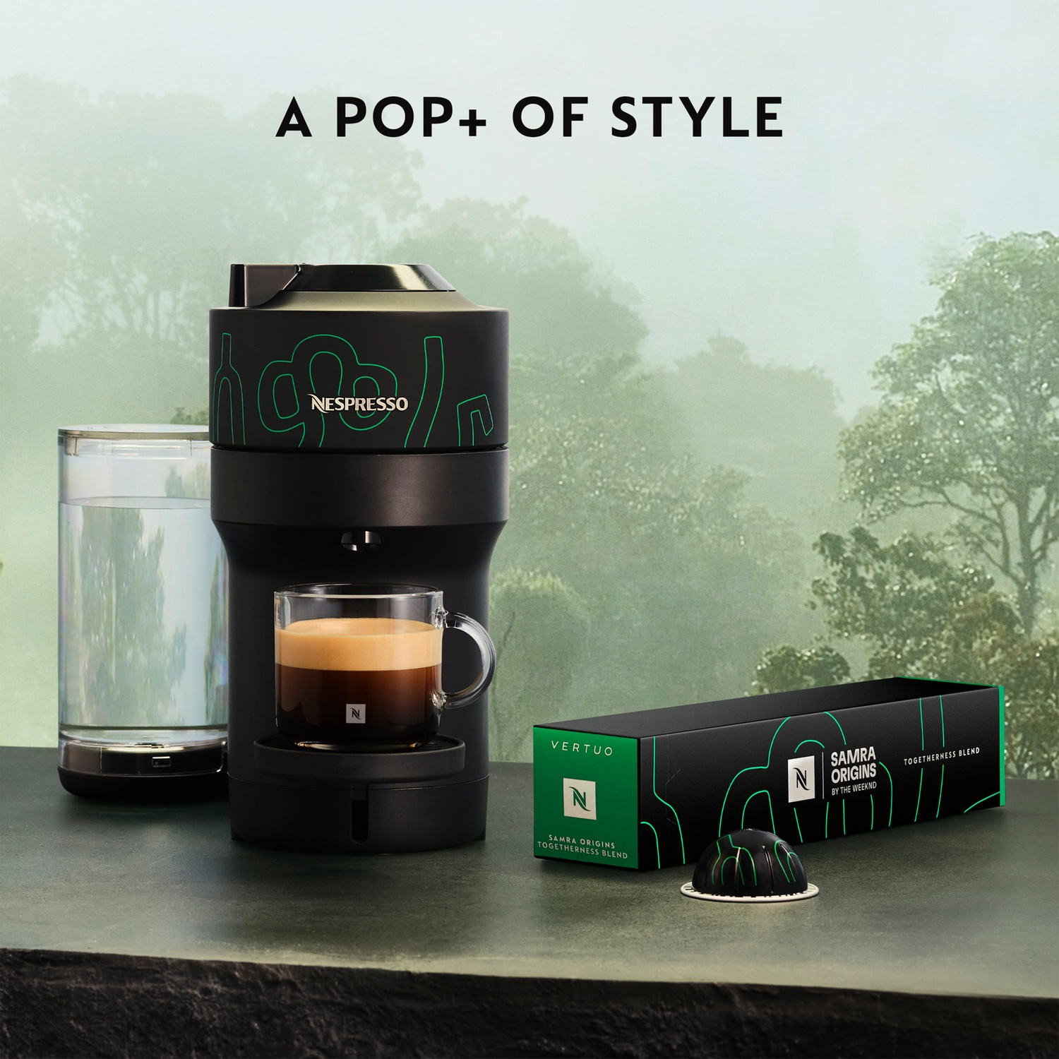 Black Nespresso machine with coffee in a glass cup, a water glass, and a Samra Origins box, all on a green surface, forest background.