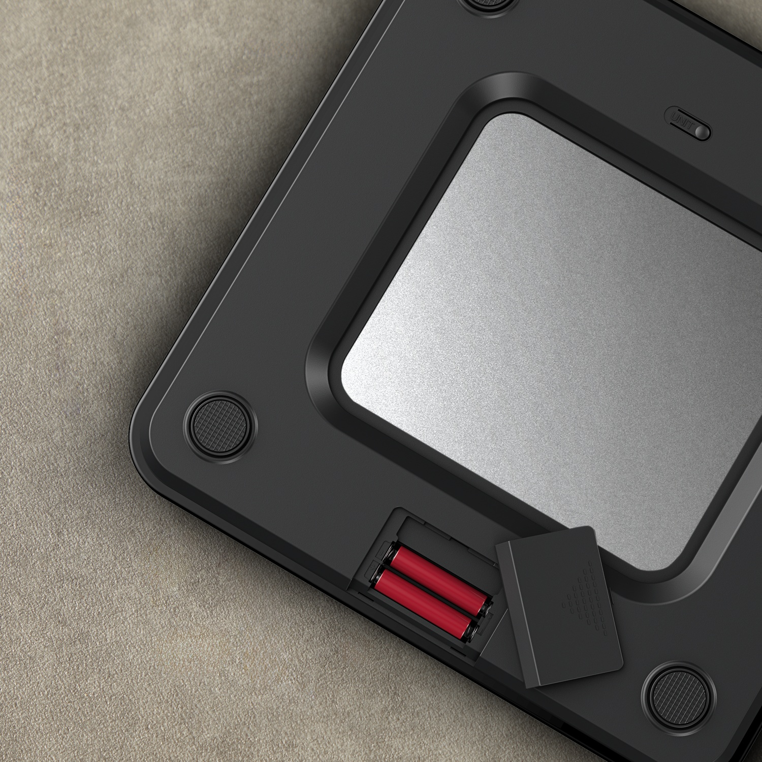 Black digital scale with silver surface. Red batteries are visible in the open battery compartment. Placed on a beige textured surface.