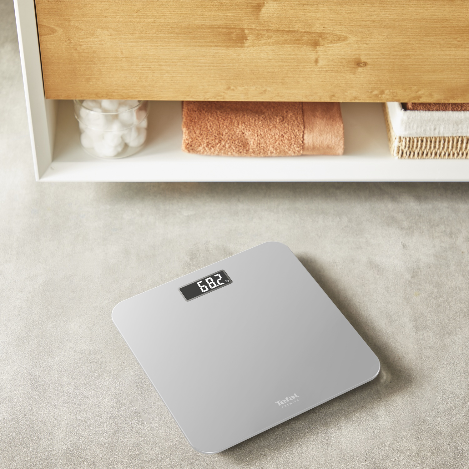 A silver scale displays 68.2 kg. A wooden cabinet with a shelf holding a towel, cotton balls, and folded towels is above.
