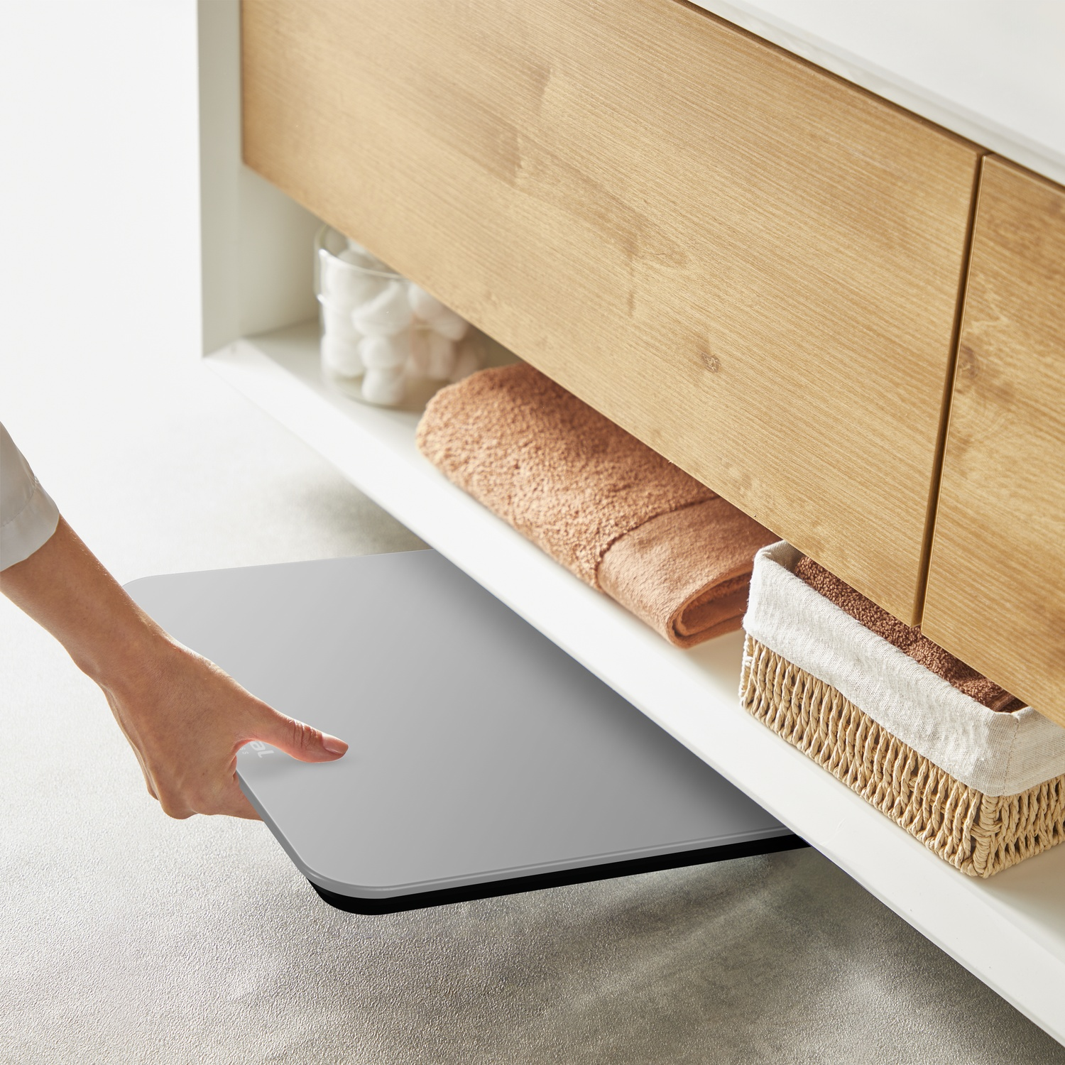 A hand pulls out a gray shelf under a wooden cabinet. A towel, basket, and jar of cotton balls are visible on the shelf.