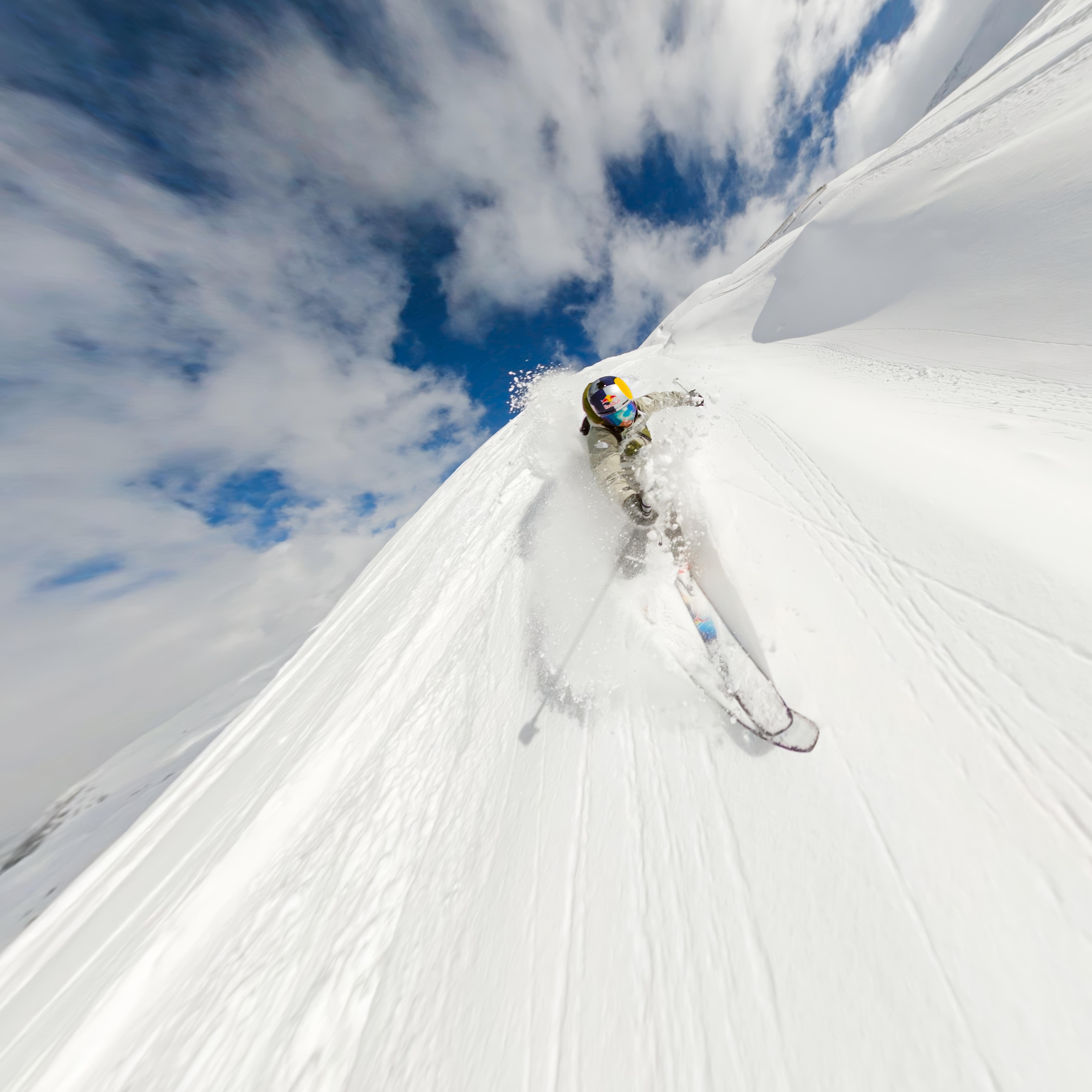 Skifahrer fährt einen schneebedeckten Berg hinunter. Blauer Himmel und Wolken im Hintergrund. Schnee spritzt um den Skifahrer herum.