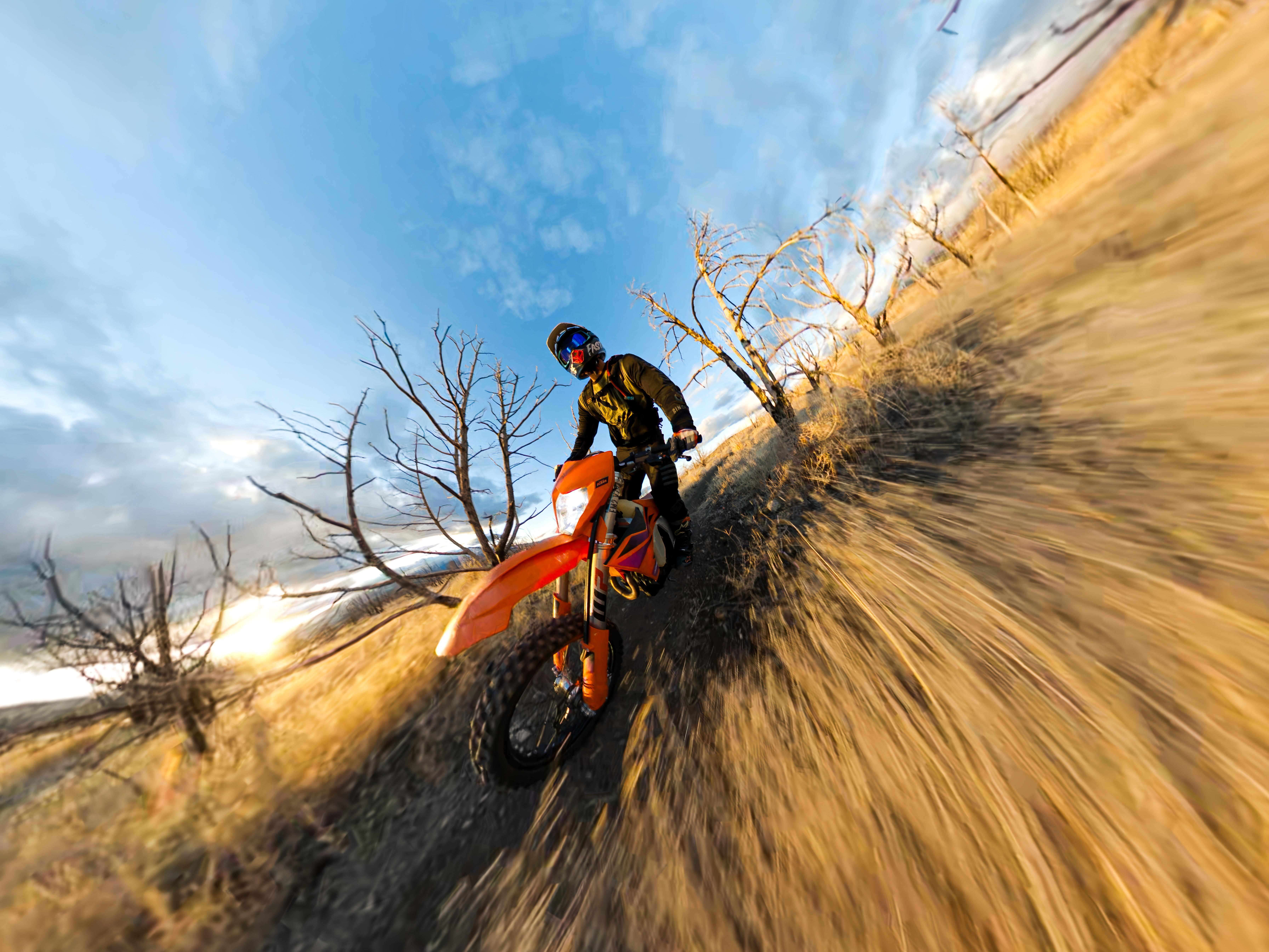 Ein Mann fährt mit einem orangen Dirtbike bergauf. Der Himmel ist blau mit Wolken. Trockenes Gras und kahle Bäume sind im Hintergrund.