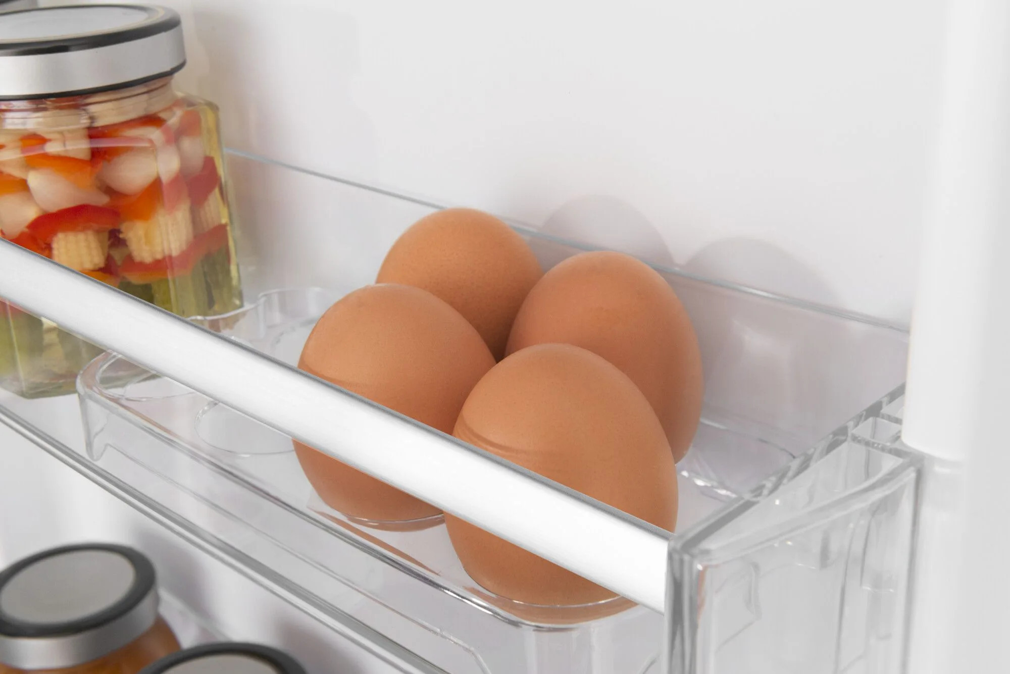 Cztery brązowe jajka w przezroczystej komorze lodówki. Słoik z marynowanymi warzywami po lewej stronie. Białe wnętrze lodówki.